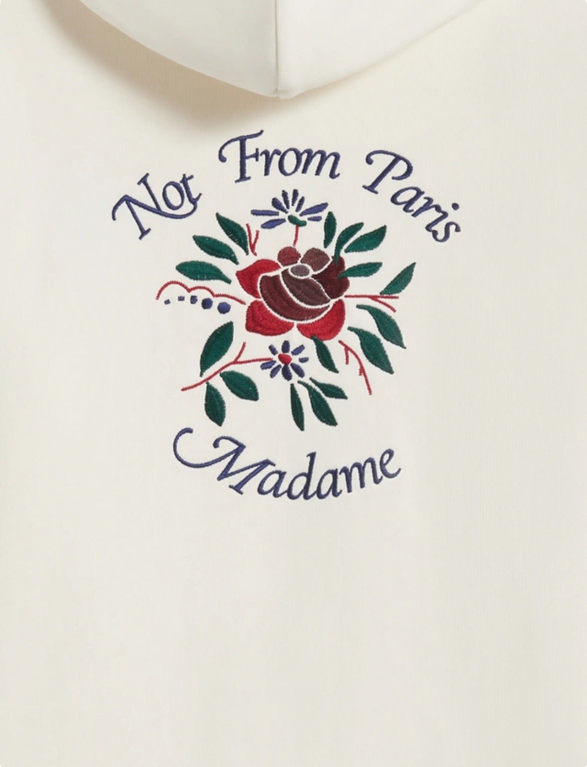 The DROLE DE MONSIEUR E-HO169-CO127-CM LE HOODIE FLEURS CREAM is a white French terry hoodie adorned with exquisite Crillon embroidery and the phrase "Not From Paris Madame" framing a vivid floral design in red, green, and purple hues. Crafted from 100% cotton for unparalleled comfort and style.