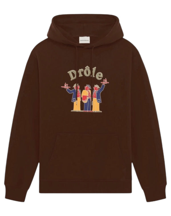 The DROLE DE MONSIEUR E-HO172-CO127-DBN LE HOODIE CRILLON BRW is a brown French terry hoodie with a front pocket, featuring an illustrated design of four figures and the word "Drôle" above them, crafted from 100% cotton.