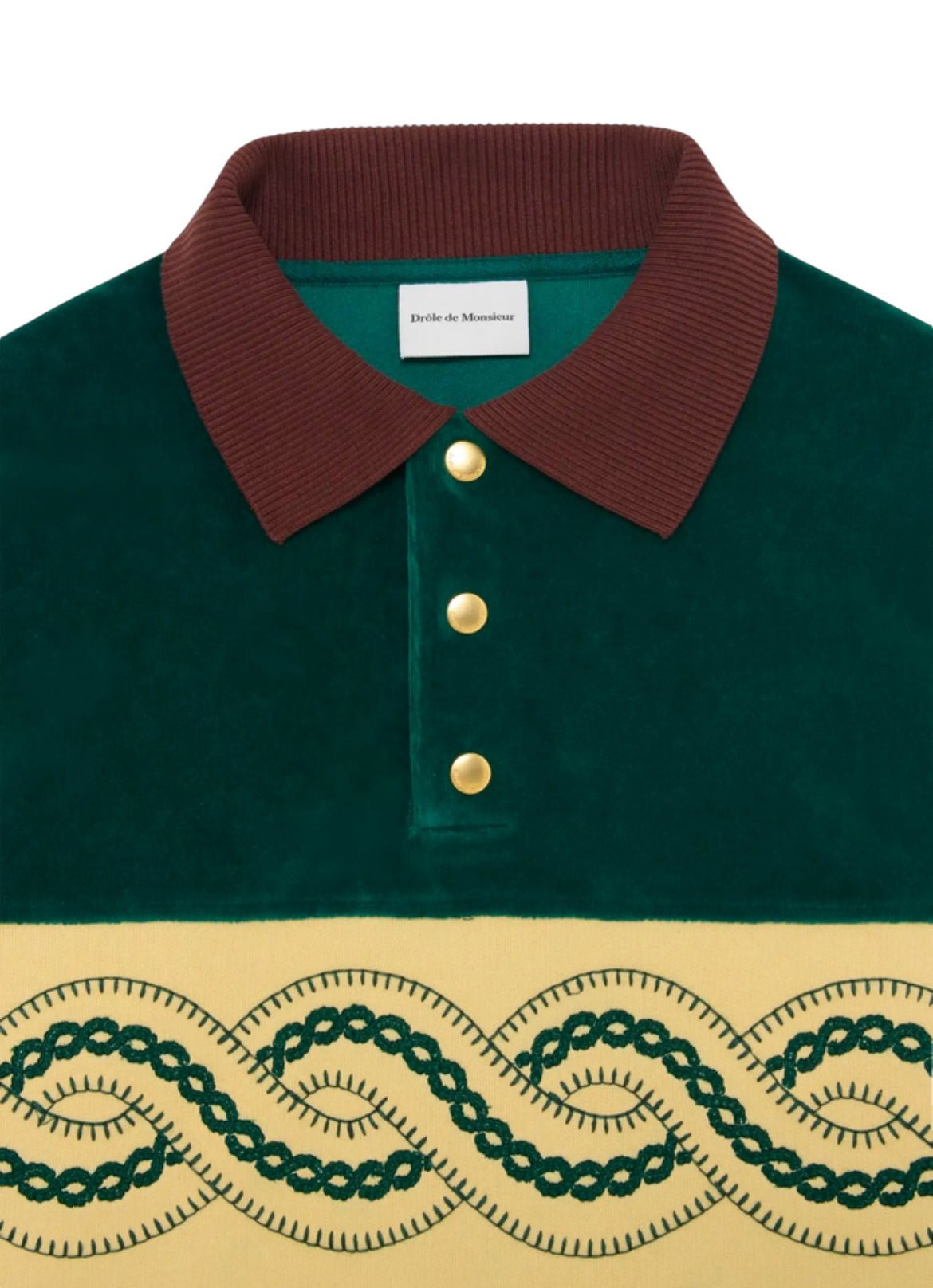 Close-up of the DROLE DE MONSIEUR E-PL133-CO171-DGN LE POLO SWEAT GRN. It features a stylish green fabric with a contrasting brown collar and elegant gold buttons. The front showcases an intricate dark green embroidered pattern on a beige section, enhanced by braided embroidery for added texture.