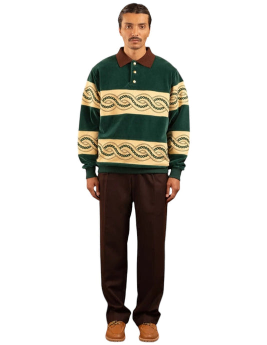 A person stands wearing the DROLE DE MONSIEUR E-PL133-CO171-DGN LE POLO SWEAT GRN, featuring green and beige stripes with braided embroidery and a brown collar, paired with brown pants and brown shoes against a plain white background.