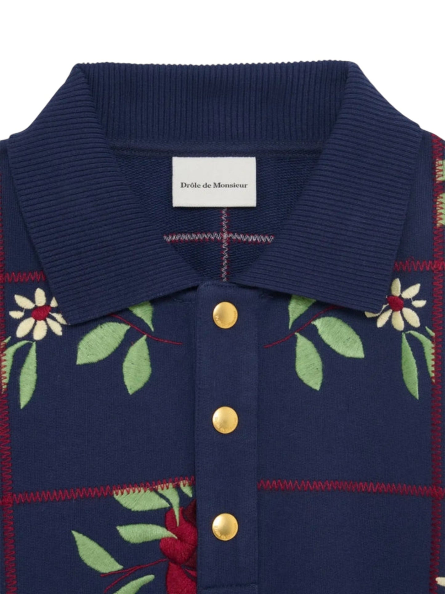 Close-up of the DROLE DE MONSIEUR E-PL135-CO127-NY LE POLO FLEURS NVY, featuring a navy blue fleece fabric with floral patterns, gold buttons, and a ribbed collar. The label reads "Drôle de Monsieur".