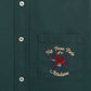 Close-up of the DROLE DE MONSIEUR E-SH189-PL LA CHEMISE SLOGAN FLEUR GRN, a dark green long-sleeved shirt with a pocket. The pocket features embroidered text reading "Not From Paris Madame" and a red and blue Slogan Fleurs floral design.