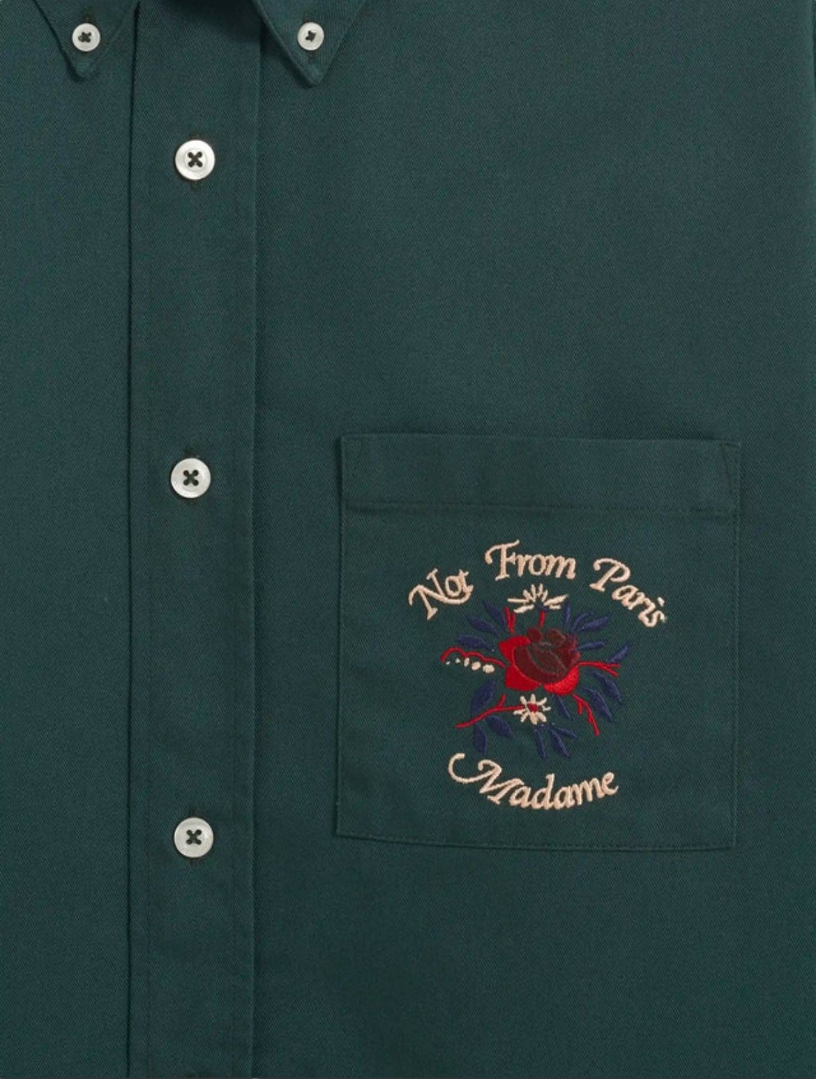 Close-up of the DROLE DE MONSIEUR E-SH189-PL LA CHEMISE SLOGAN FLEUR GRN, a dark green long-sleeved shirt with a pocket. The pocket features embroidered text reading "Not From Paris Madame" and a red and blue Slogan Fleurs floral design.