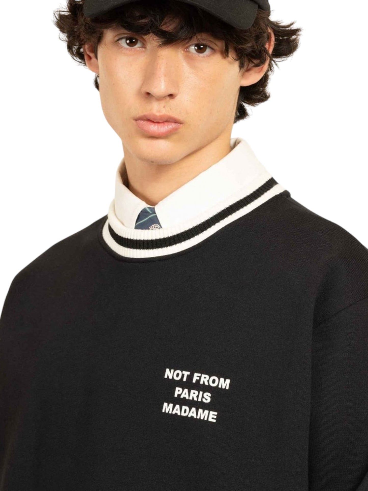 Someone wearing the Drôle de Monsieur F-SW140-CO127-Bl Le Sweatshirt Black with "Not From Paris Madame" text over a collared shirt and tie, along with a black cap.