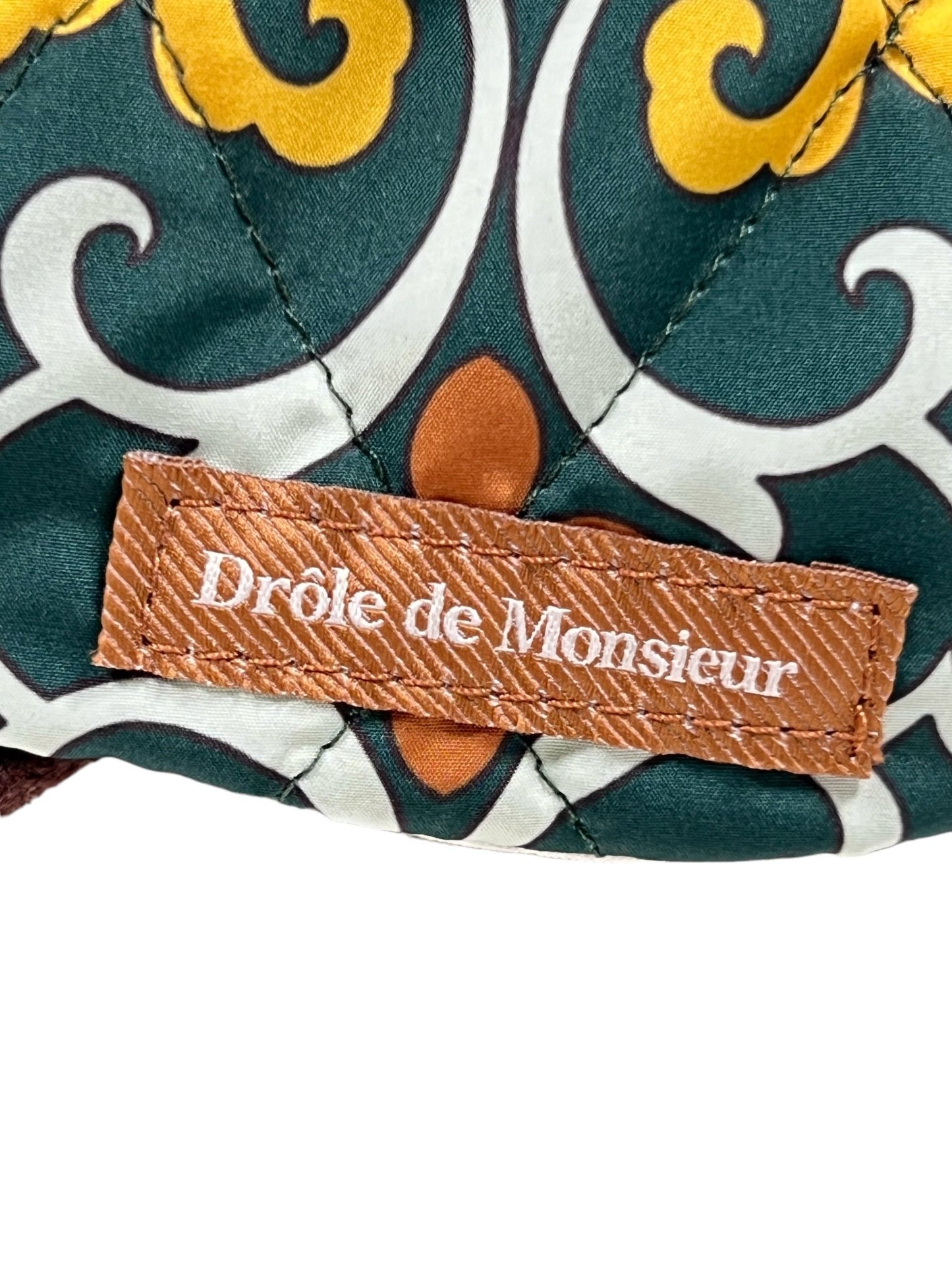 Close-up of a digitally printed patterned fabric with green, yellow, and brown elements, featuring a brown label that reads "DROLE DE MONSIEUR" from the DROLE DE MONSIEUR HAT B-CP123 LA CASQUETTE FAIENCE GREEN.