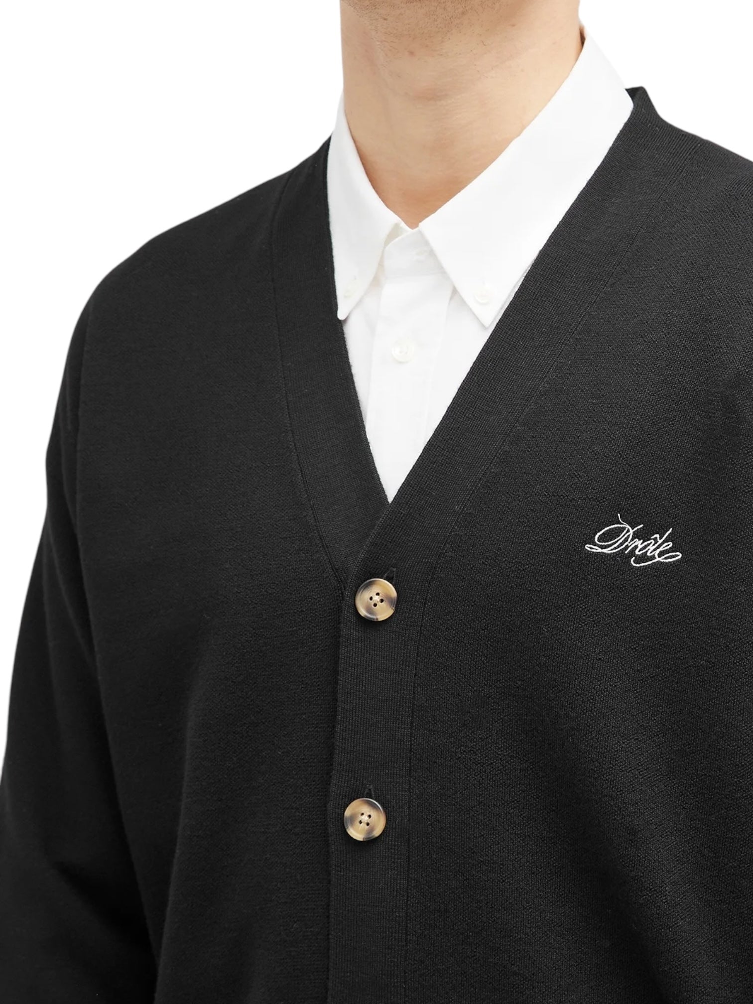 Someone is wearing the Drôle de Monsieur Perm-CA123-WO007-BL LE Cardigan Blk, a black wool cardigan with ribbed trim and buttons, layered over a white collared shirt. It features a small Drôle embroidery logo on the left side.