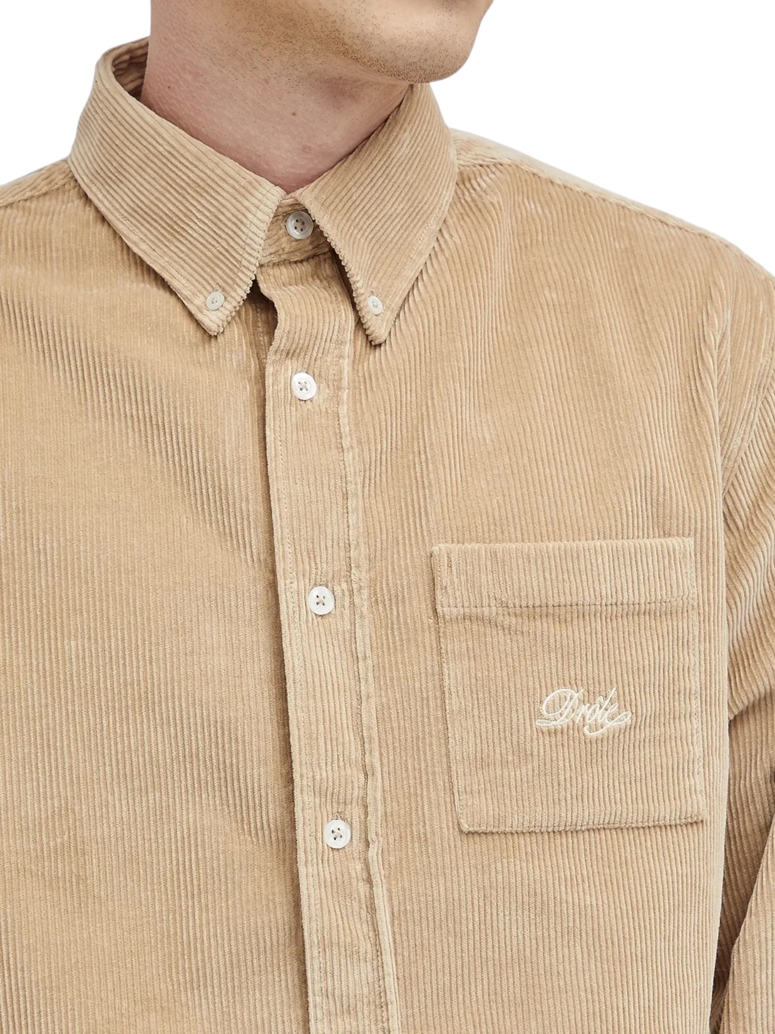 A person wearing the Drôle de Monsieur La Chemise Beige, featuring a corduroy button-up design with embroidery on the pocket and long sleeves.