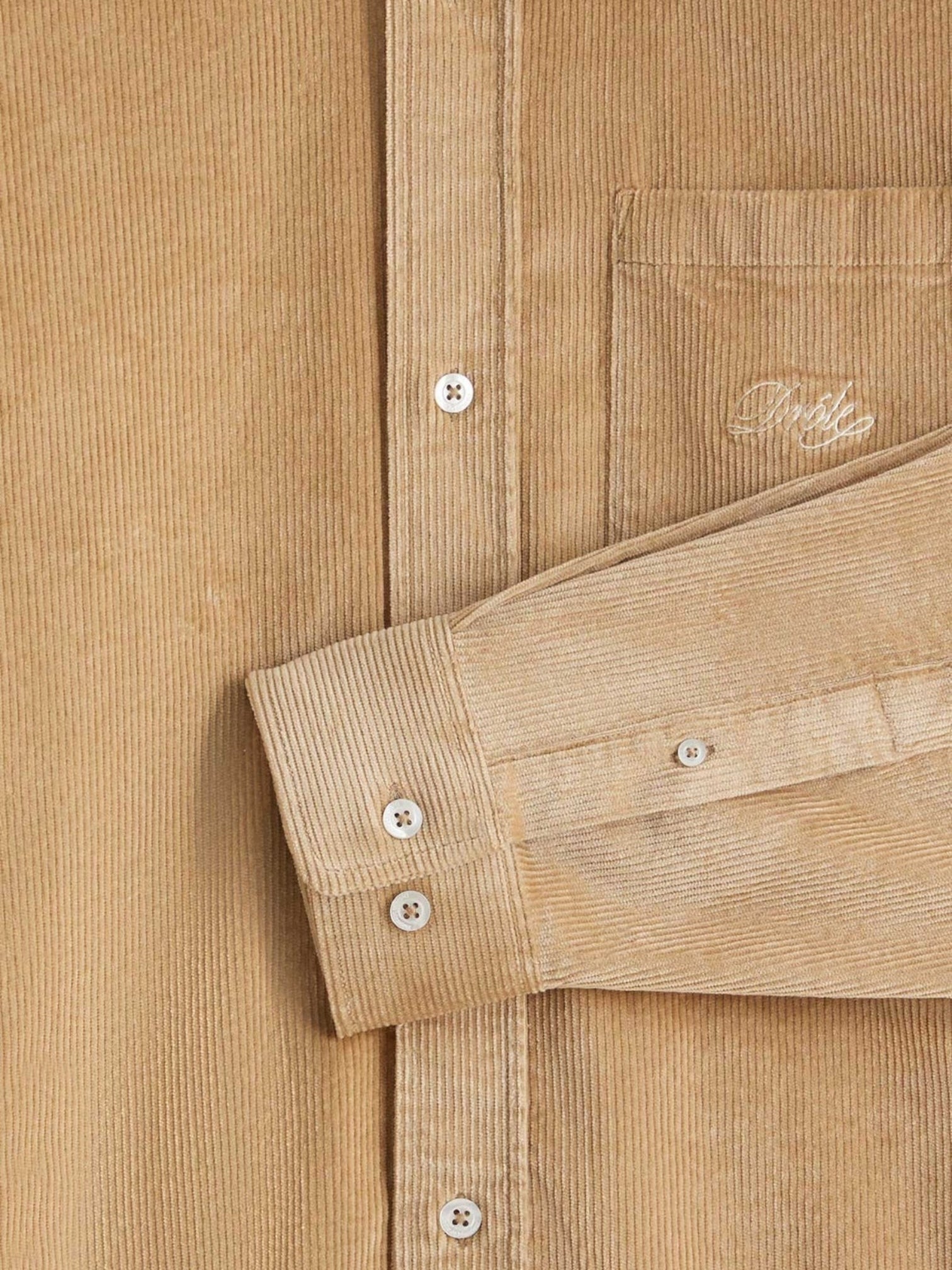 Close-up of the beige Drôle de Monsieur Perm-SH172-CO156-BG La Chemise, showcasing embroidery on the breast pocket and buttoned long sleeves.