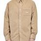 A person wears the Drôle de Monsieur Perm-SH172-CO156-BG La Chemise Beige long-sleeve corduroy shirt with embroidery on the breast pocket, standing against a white background.