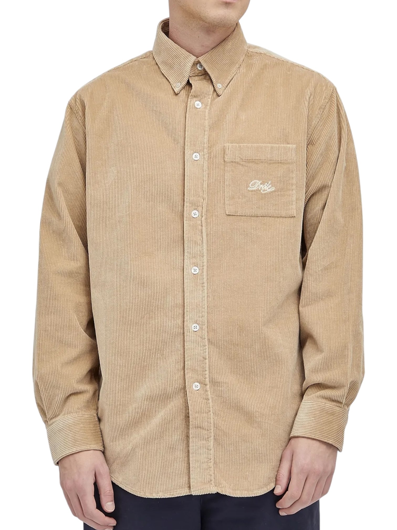A person wears the Drôle de Monsieur Perm-SH172-CO156-BG La Chemise Beige long-sleeve corduroy shirt with embroidery on the breast pocket, standing against a white background.