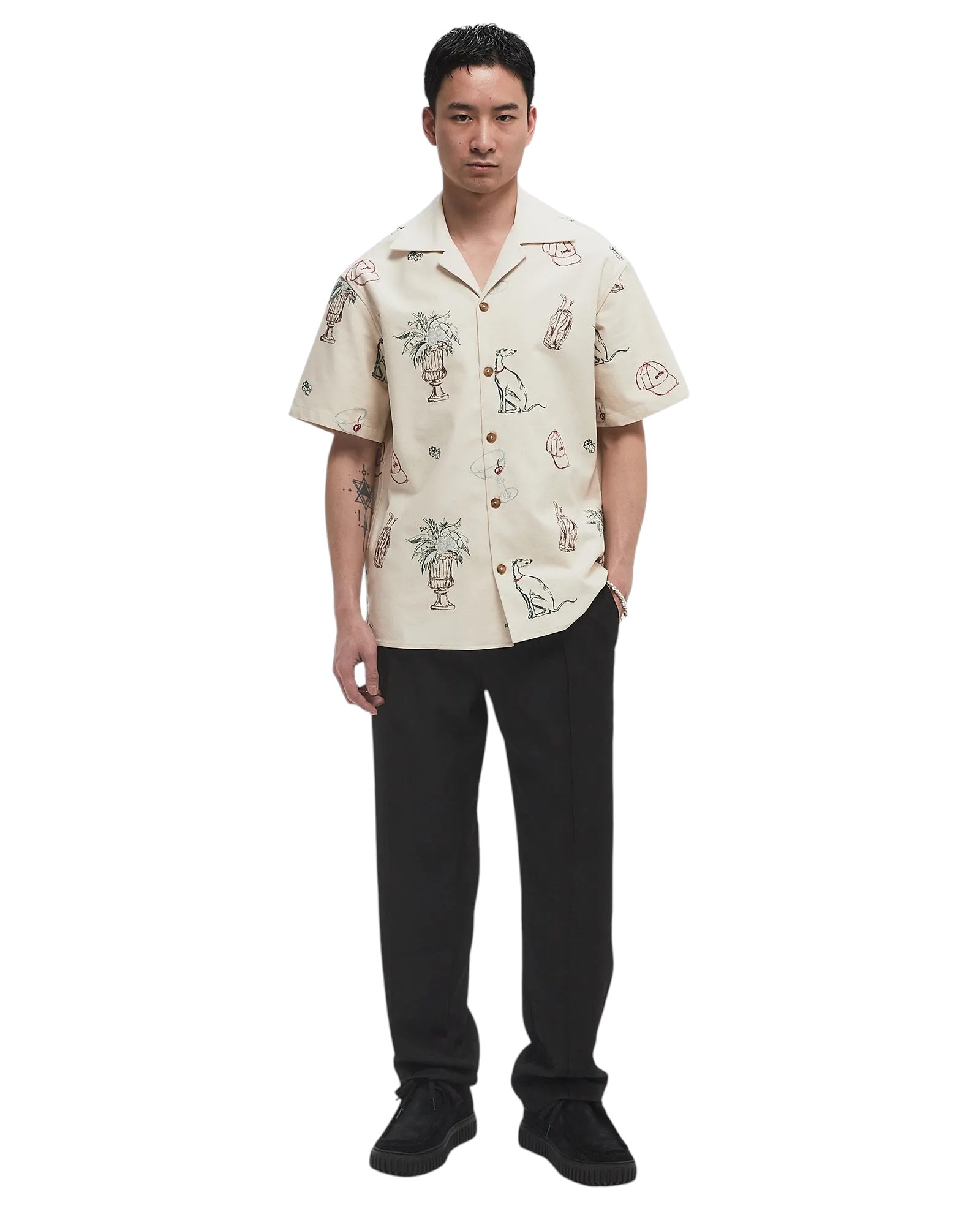 A man faces forward wearing the Drôle de Monsieur SH214-CO198-EC La Chemise Brodee Ecru, a short-sleeved, cream cotton-blend shirt with abstract line drawings, paired with black pants and black shoes.