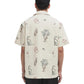 A man seen from the back wears the Drôle de Monsieur SH214-CO198-EC La Chemise Brodee Ecru, a light-colored cotton blend short-sleeved shirt with embroidered dogs, plants, martini glasses, and bags.