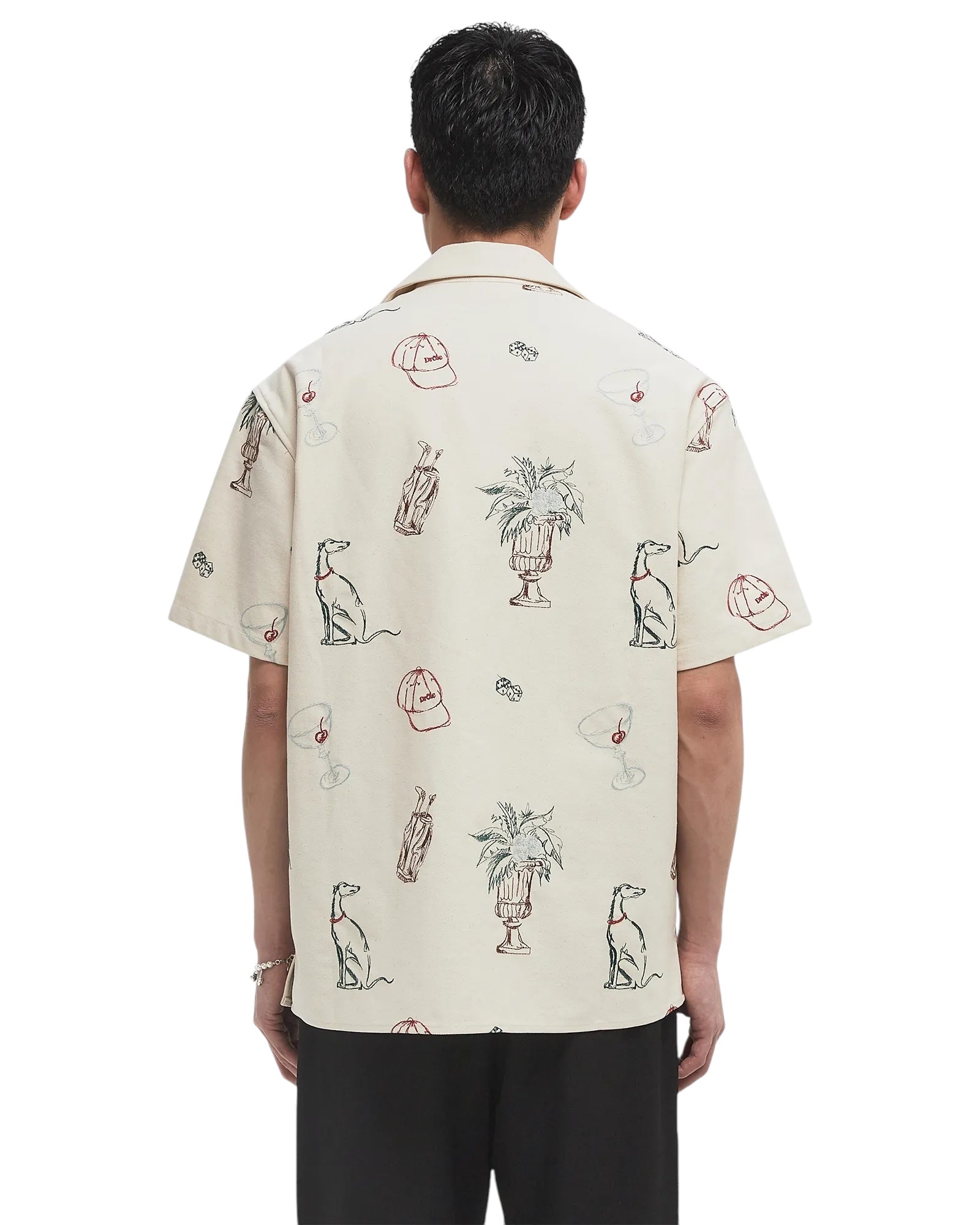 A man seen from the back wears the Drôle de Monsieur SH214-CO198-EC La Chemise Brodee Ecru, a light-colored cotton blend short-sleeved shirt with embroidered dogs, plants, martini glasses, and bags.