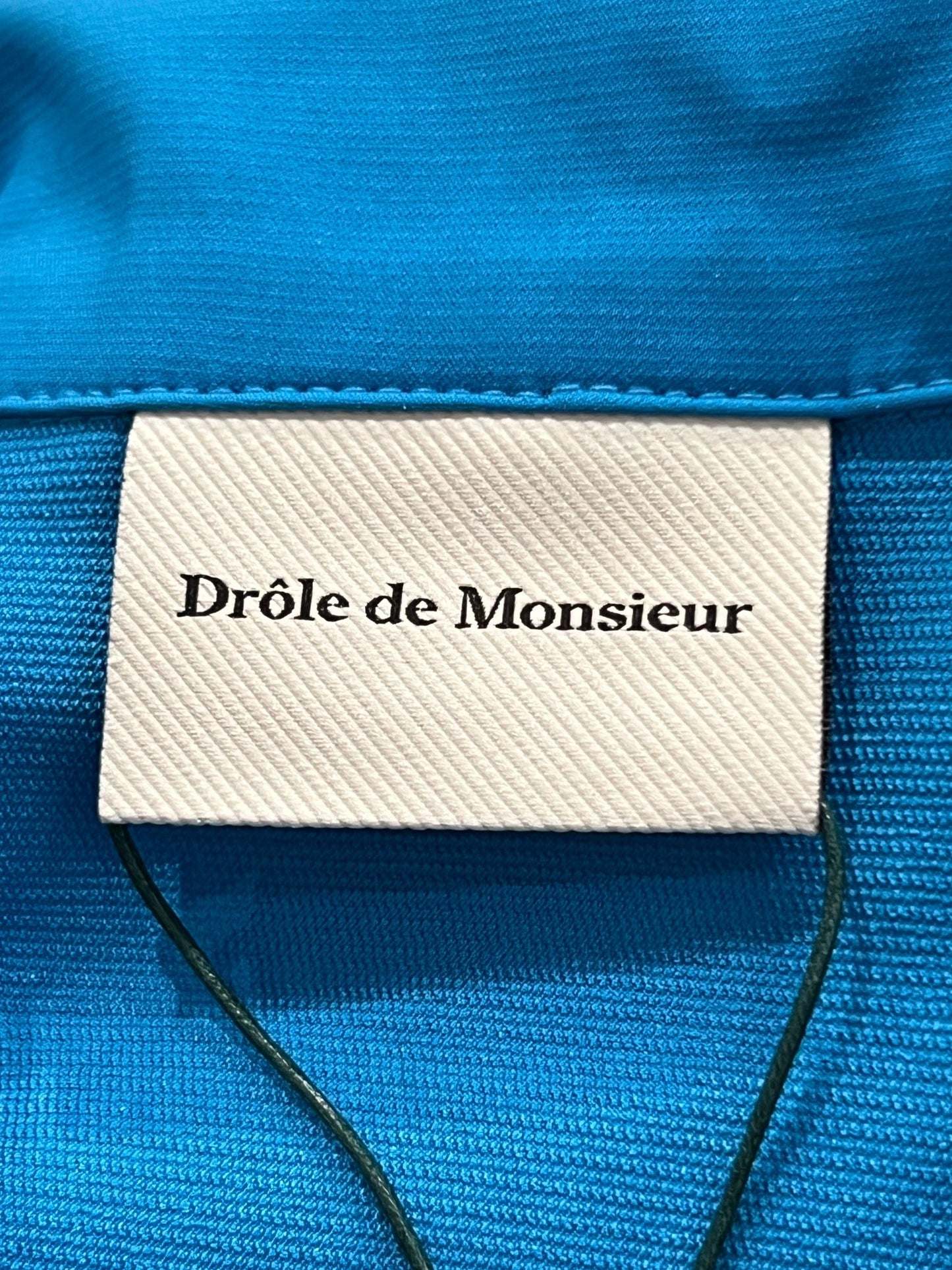 Clothing label reading "DROLE DE MONSIEUR SHIRT D-JT174-PL007-BE LA VESTE SLOGAN" on a technical fabric background.