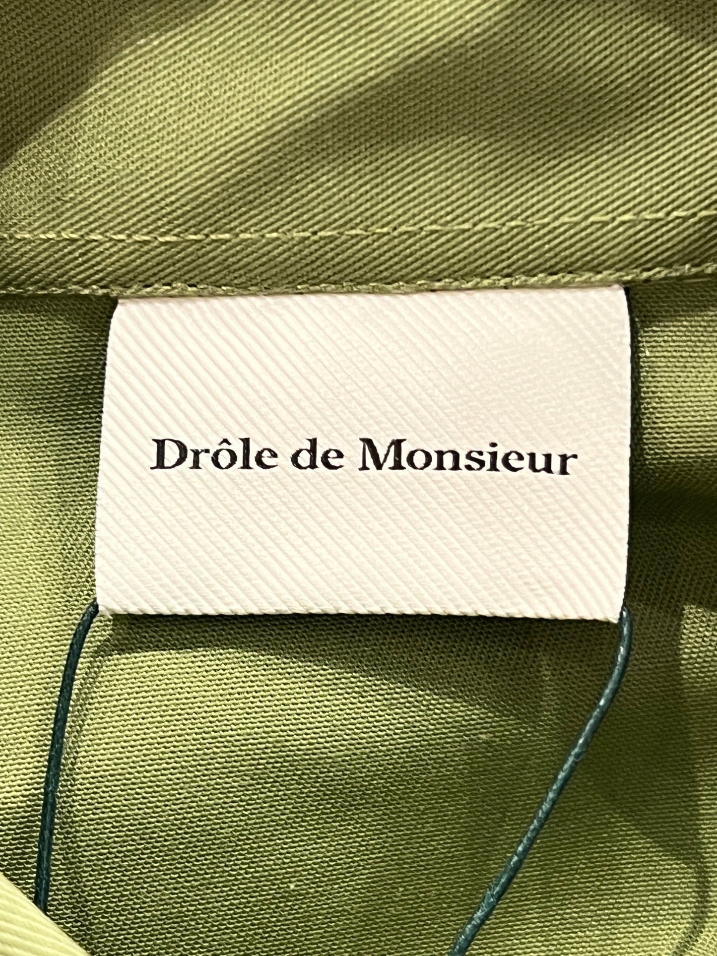 Clothing label with the DROLE DE MONSIEUR SHIRT D-SH128-PL004 LA CHEMISE SLOGAN LKK shirt stitched onto a khaki cotton blend shirt.