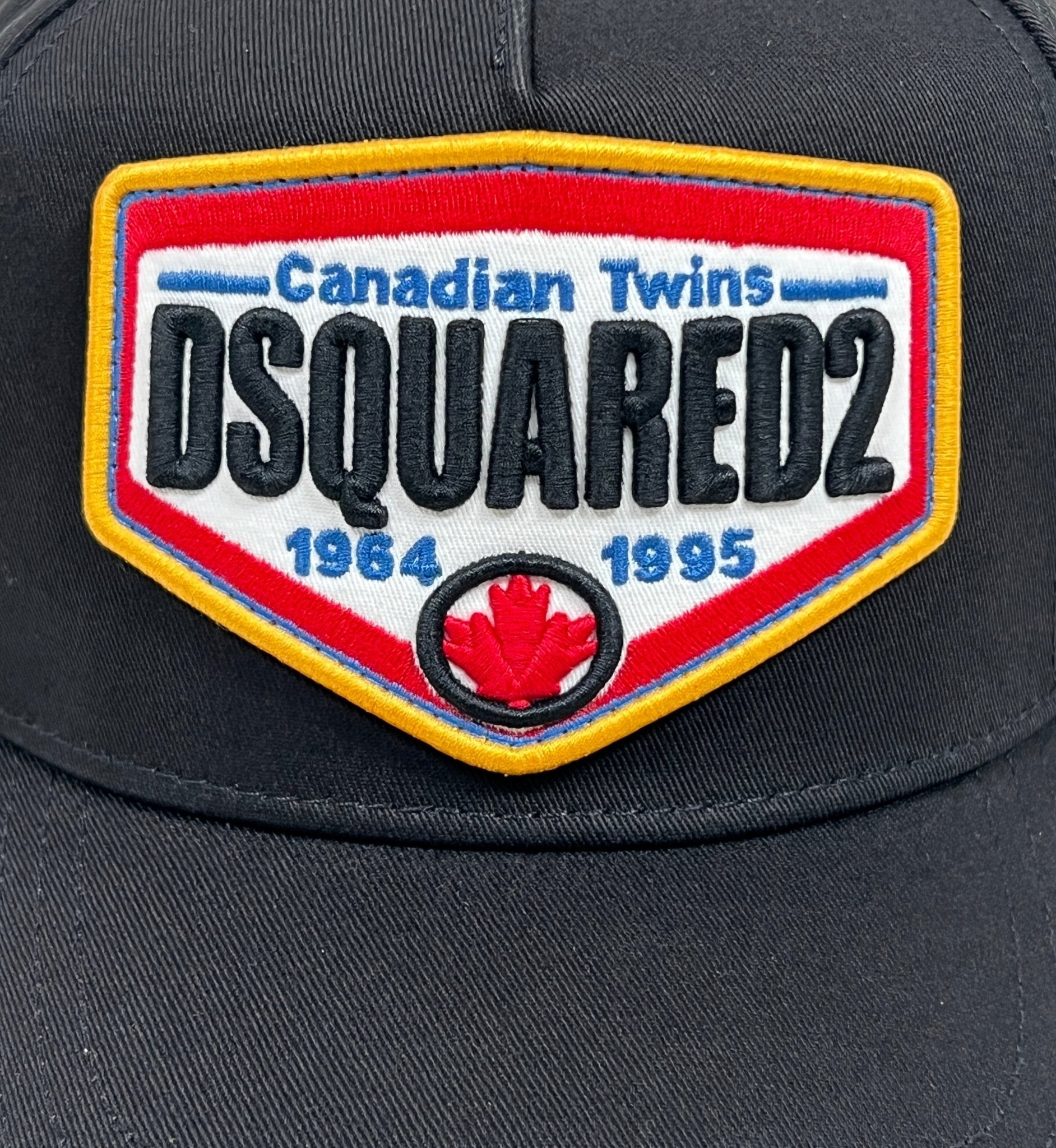 The DSQUARED2 BCM0763 Logo Baseball Cap in color 2124, by the brand DSQUARED2, is a black cap featuring a vibrant patch that reads "Canadian Twins DSQUARED2" and "1964 1995," with a prominent red maple leaf emblem at the bottom center. The adjustable strap ensures it fits perfectly on any head size.