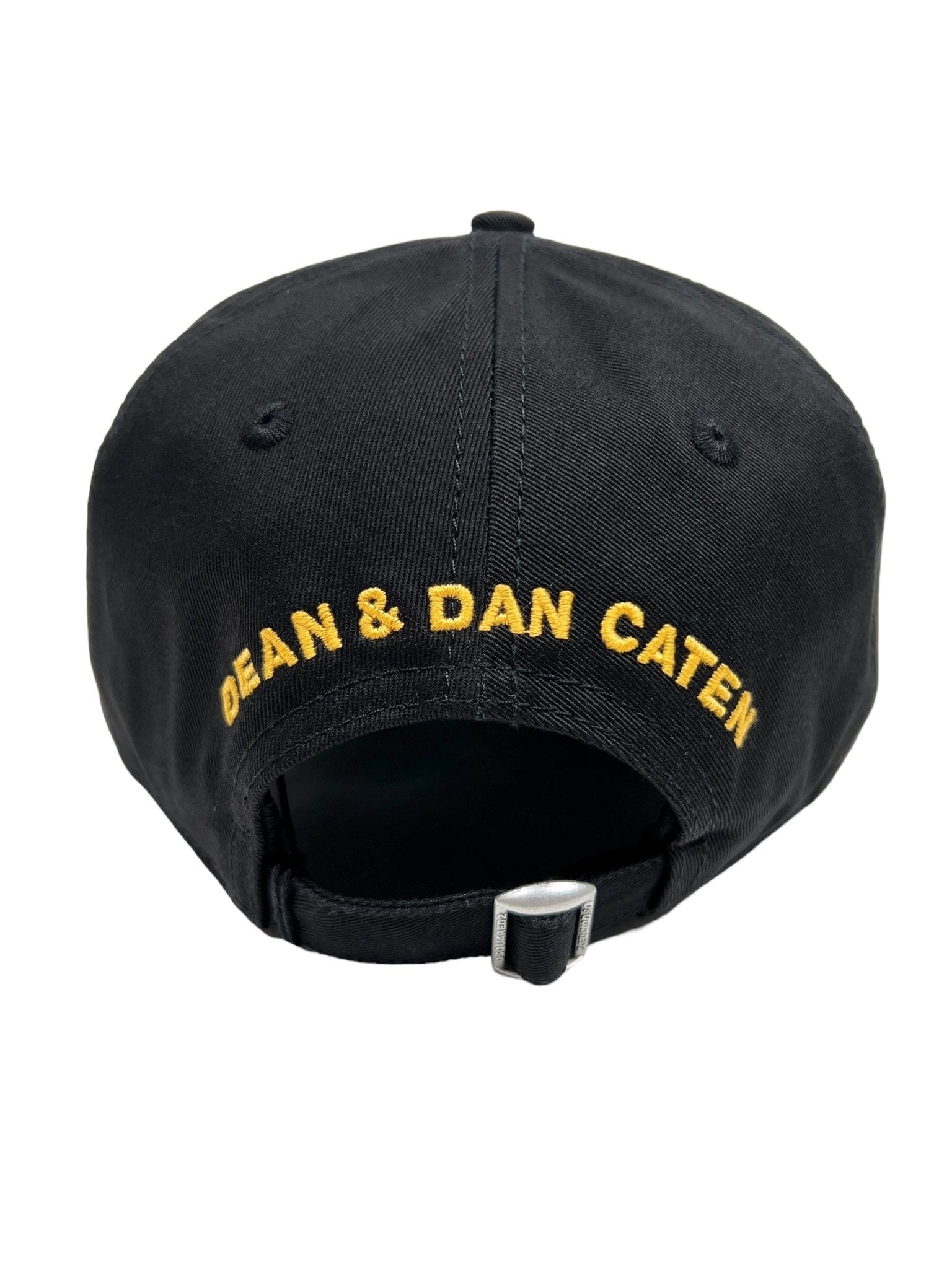 The image shows the back view of a DSQUARED2 BCM0763 LOGO BASEBALL CAP COL.2124 in black, featuring an adjustable strap. The text "DEAN & DAN CATEN" is embroidered in yellow, highlighting its connection to the DSQUARED2 brand.