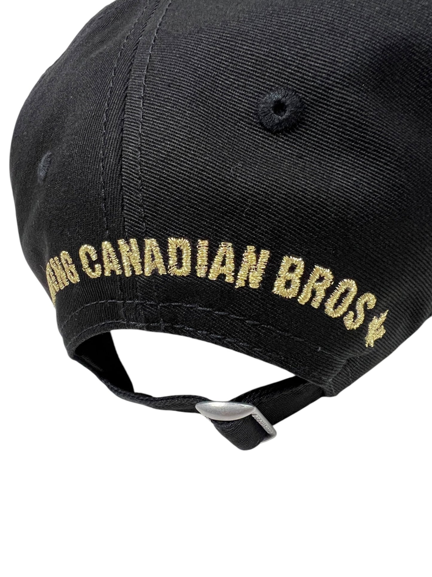 The DSQUARED2 BCM0903 Baseball Cap Gabardine-Nero is a stylish cotton gabardine cap featuring gold embroidered "BIG CANADIAN BROS" on the back, with a metal buckle strap for easy adjustment.