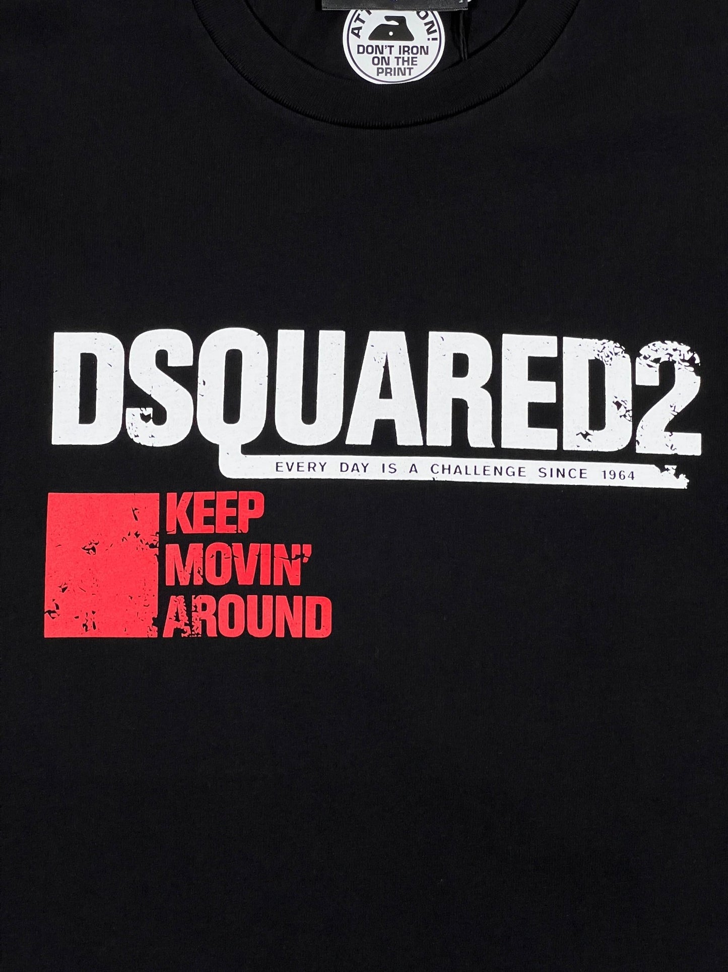 The DSQUARED2 S71GD1462 Cool Fit Tee in black features white "DSQUARED2" text, a tagline "Every Day is a Challenge Since 1964," and a red box with the text "Keep Movin' Around." The tag advises, "Don't Iron Print." Made from 100% cotton, this men's stylish T-shirt from DSQUARED2 seamlessly blends comfort and fashion.