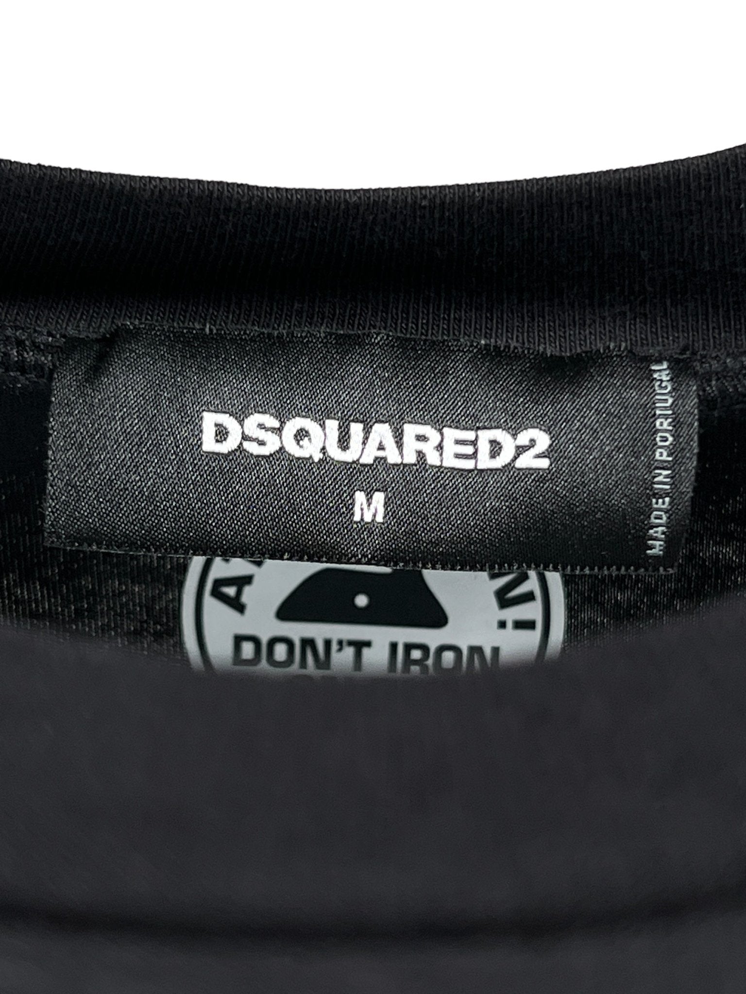 Close-up of a clothing label on a DSQUARED2 S74Gd1307 Cool Fit Tee Black in size "M," featuring the brand name "DSQUARED2." The care label below indicates it is made in Portugal and advises, "Don't Iron.