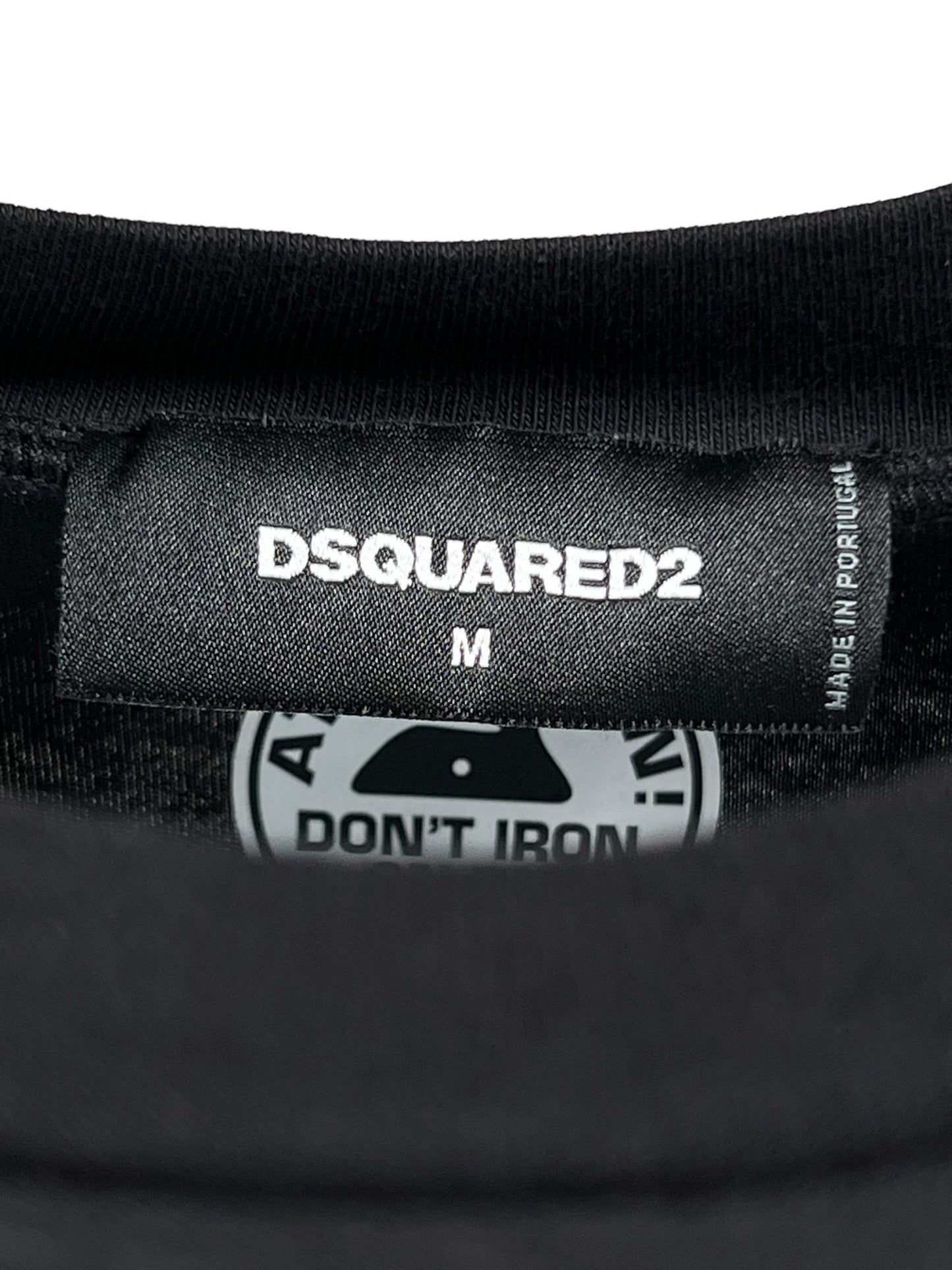 Close-up of a black label on the Dsquared2 S74Gd1316 Regular Fit Tee, size M. Made from 100% cotton, it includes washing instructions "DON'T IRON" accompanied by a warning symbol. Crafted in Portugal.