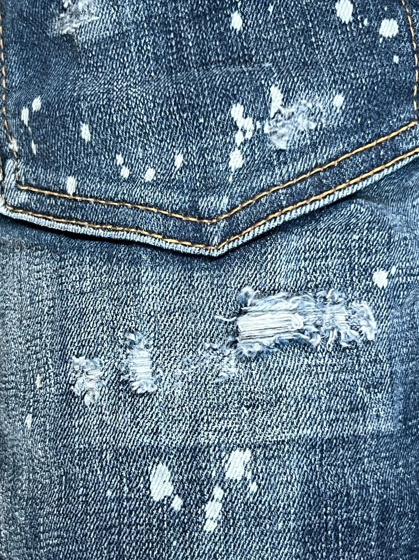 Close-up of blue denim fabric with noticeable white paint splatters and distressed, torn areas, including a pocket with frayed edges and exposed threads. This trendy ripped detailing is reminiscent of the DSQUARED2 S74LB1556 SKATER JEANS NAVY BLUE, featuring a stylish slim fit.