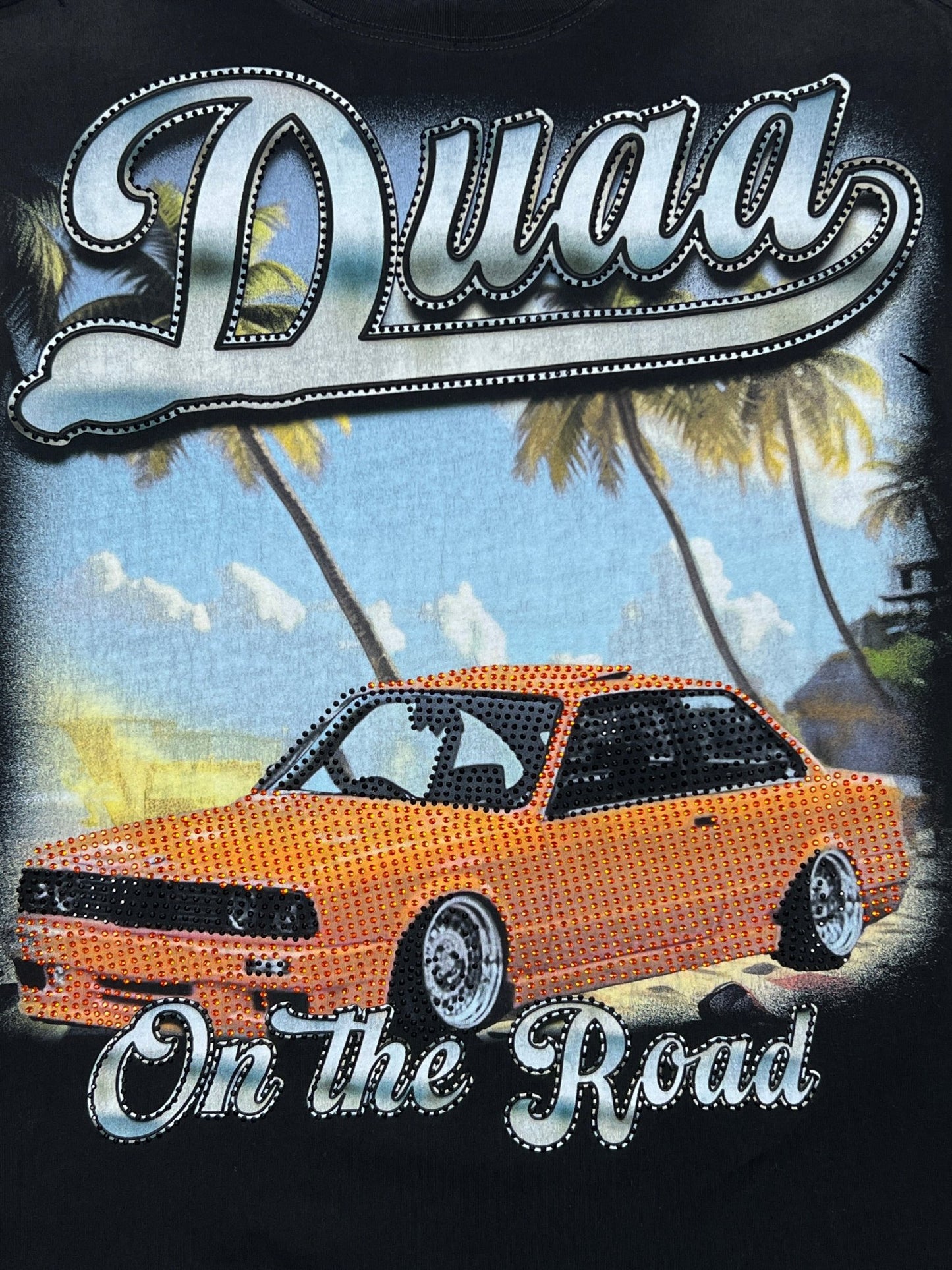 An illustration on the DUAA Duaa Destination Blk Tee Shirt shows an orange car with crystal details, palm trees, and a beach. "Duaa" and "On the Road" are in bold script above and below—perfect for your favorite black tee.