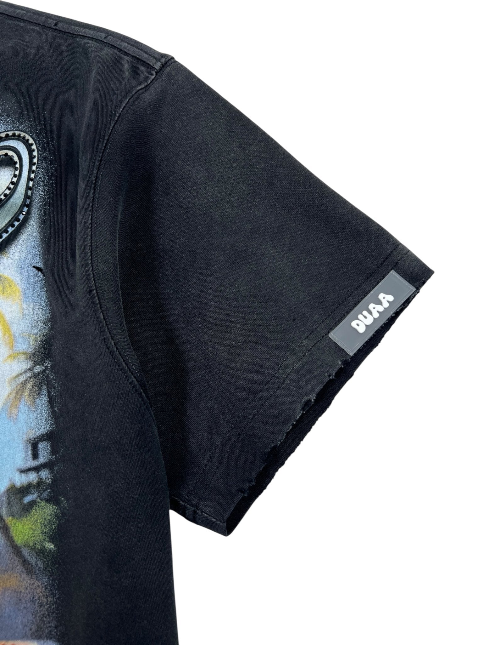 Close-up of the Duaa Destination Blk Tee Shirt by DUAA, a black short-sleeve graphic tee with a partially visible airbrushed design and a small "DUAA" label sewn onto the sleeve hem.
