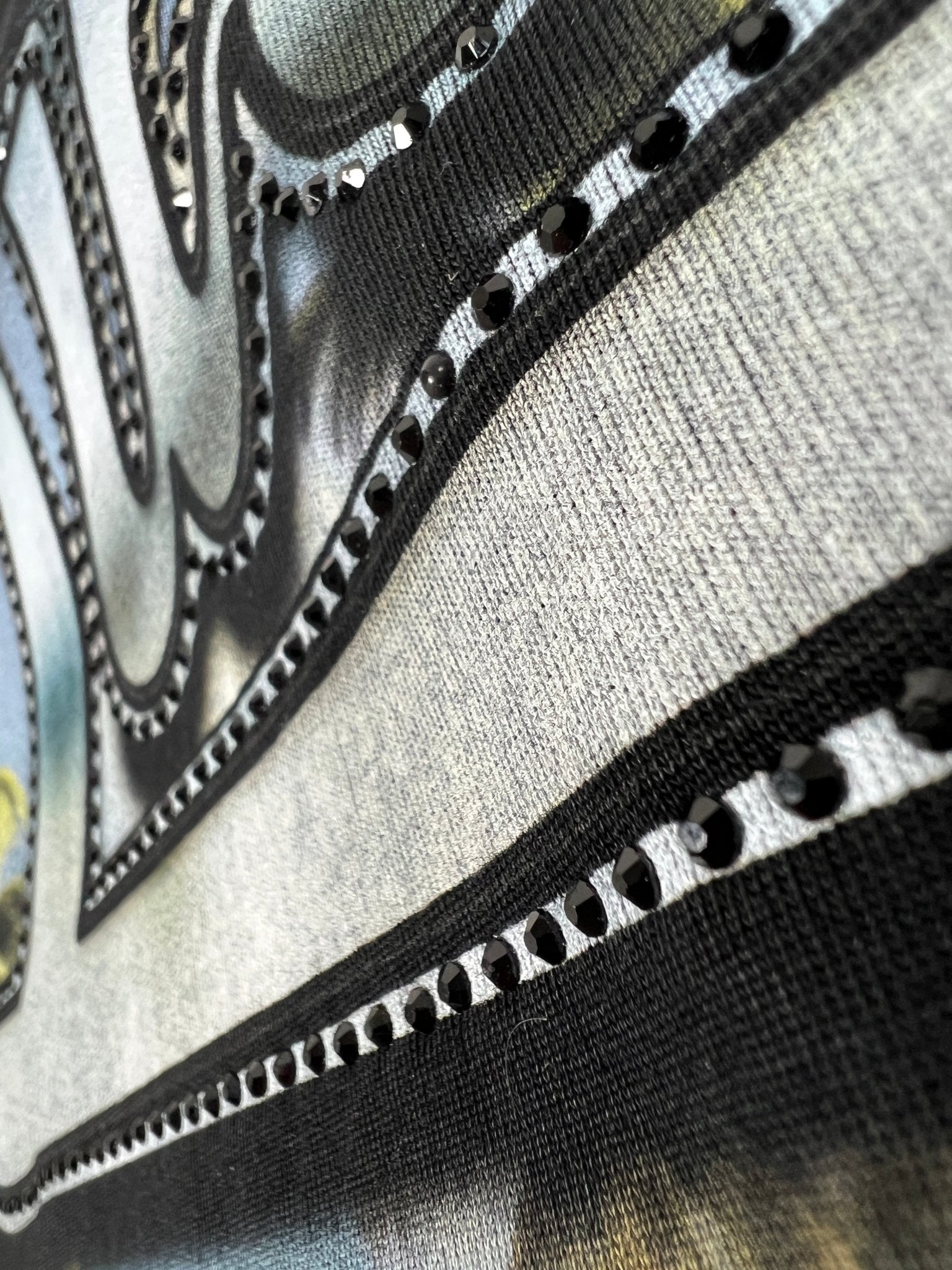 Close-up of the Duaa Destination Blk Tee Shirt by DUAA, featuring textured fabric with black lines, metallic studs, and crystal details on a gradient background that highlights detailed stitching and decorative elements.