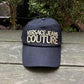 A black VERSACE E72GAZK10-EZG010-EG89 hat with the logo "Versace Jeans Couture" embroidered on the front, displayed on a wooden surface with greenery in the background.