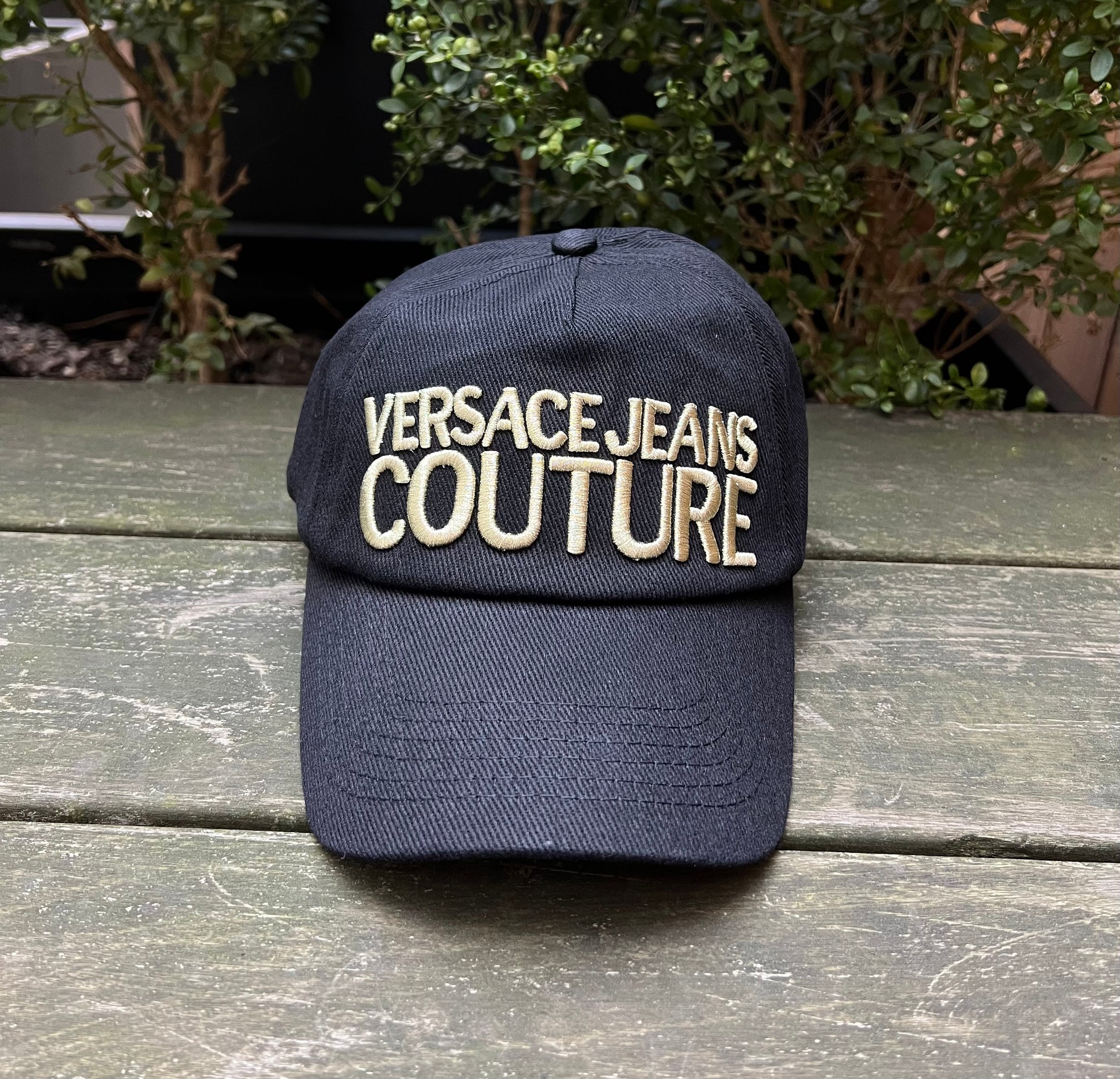 A black VERSACE E72GAZK10-EZG010-EG89 hat with the logo "Versace Jeans Couture" embroidered on the front, displayed on a wooden surface with greenery in the background.