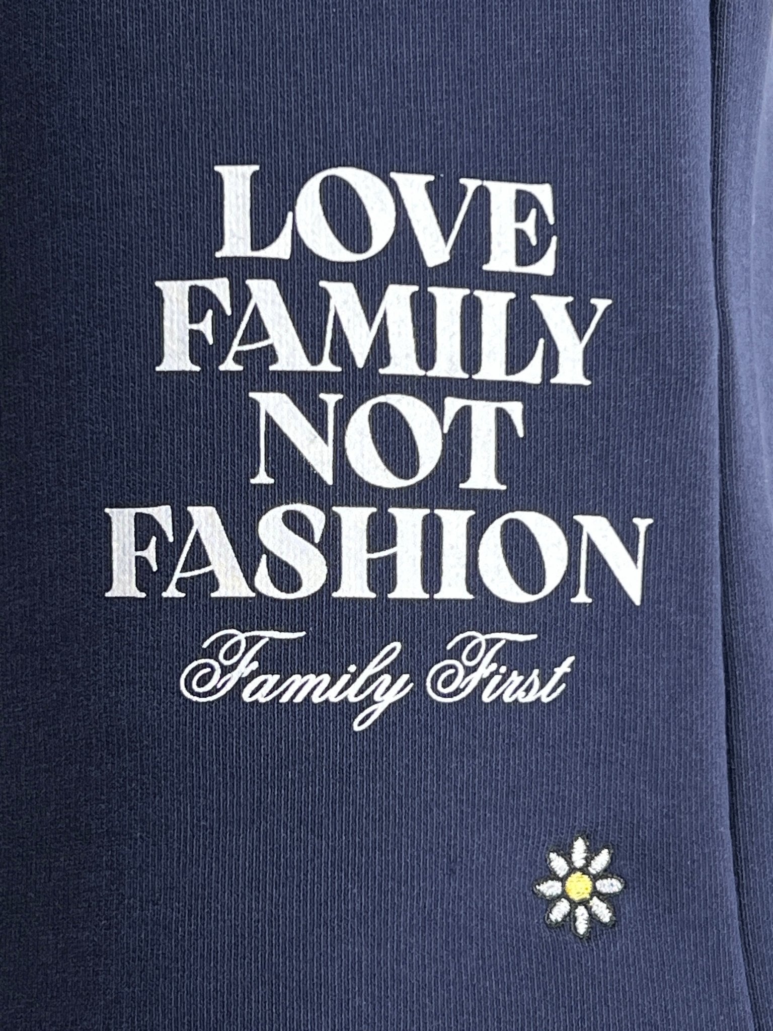 The FAMILY FIRST JOSS2503 Short Flower Dark Blue features navy cotton fabric, white "LOVE FAMILY NOT FASHION Family First" text, and a small embroidered white and yellow daisy at the bottom.