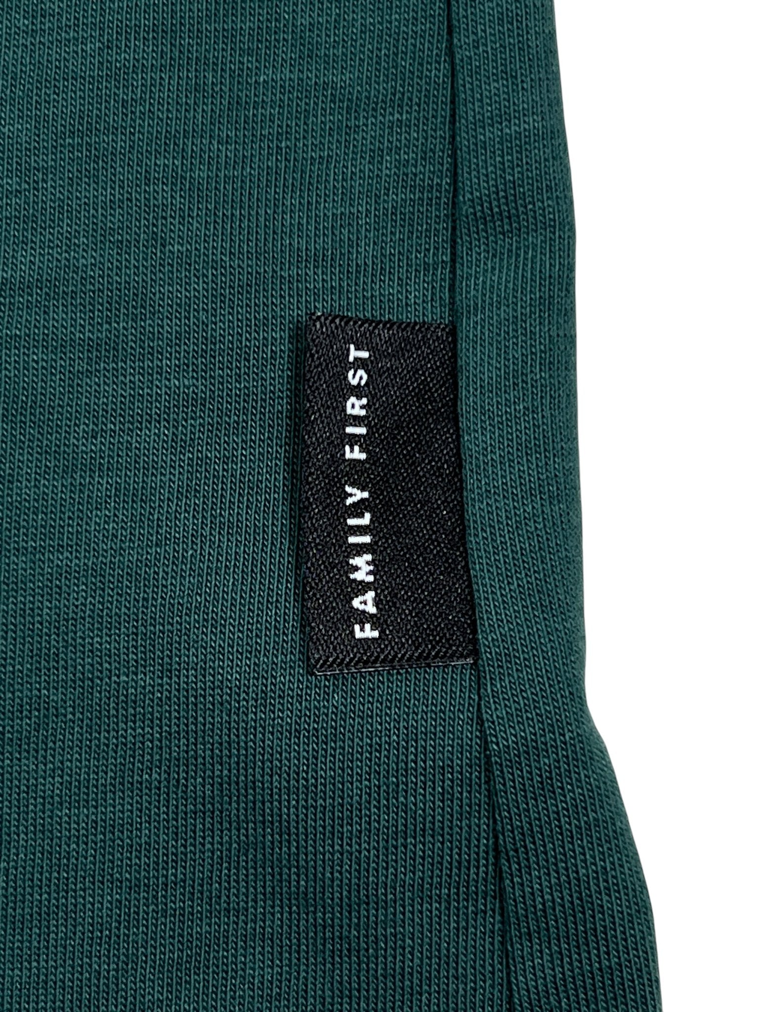 A close-up of the luxurious green ribbed fabric of the Family First TF2409GR T-shirt Symbol Green highlights a sophisticated black label with "FAMILY FIRST" in white text.