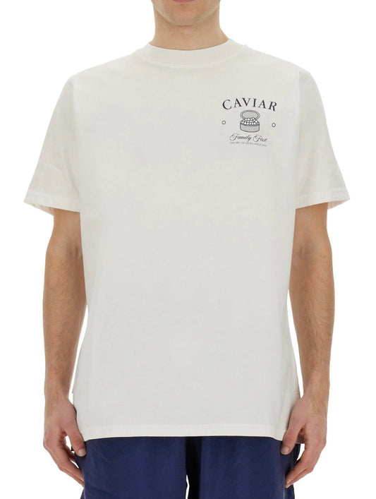 A man wearing a FAMILY FIRST TS2404 T-SHIRT CAVIAR WHT with "caviar" and decorative elements printed in gray near the upper back.