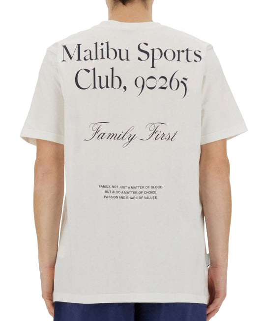 Rear view of a person wearing a white FAMILY FIRST graphic t-shirt with "malibu sports club, 90265" and "family first" printed in black text, along with a definition of family.