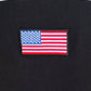 A close-up showcases the embroidered American flag patch on black fabric, with partial white lettering above, capturing the essence of Family First. This design is ideal for the Family First TS2416 College Black T-Shirt, made from 100% cotton.