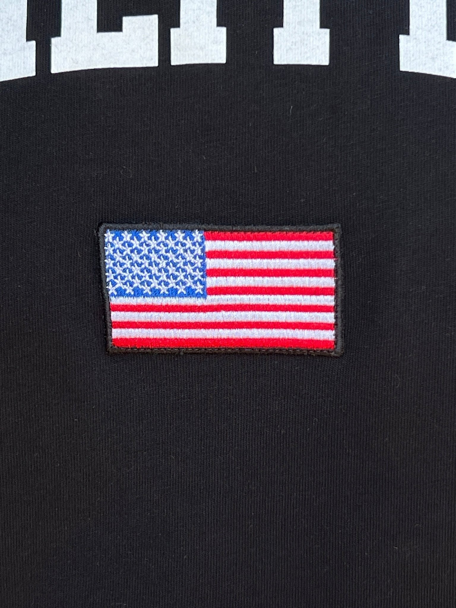 A close-up showcases the embroidered American flag patch on black fabric, with partial white lettering above, capturing the essence of Family First. This design is ideal for the Family First TS2416 College Black T-Shirt, made from 100% cotton.