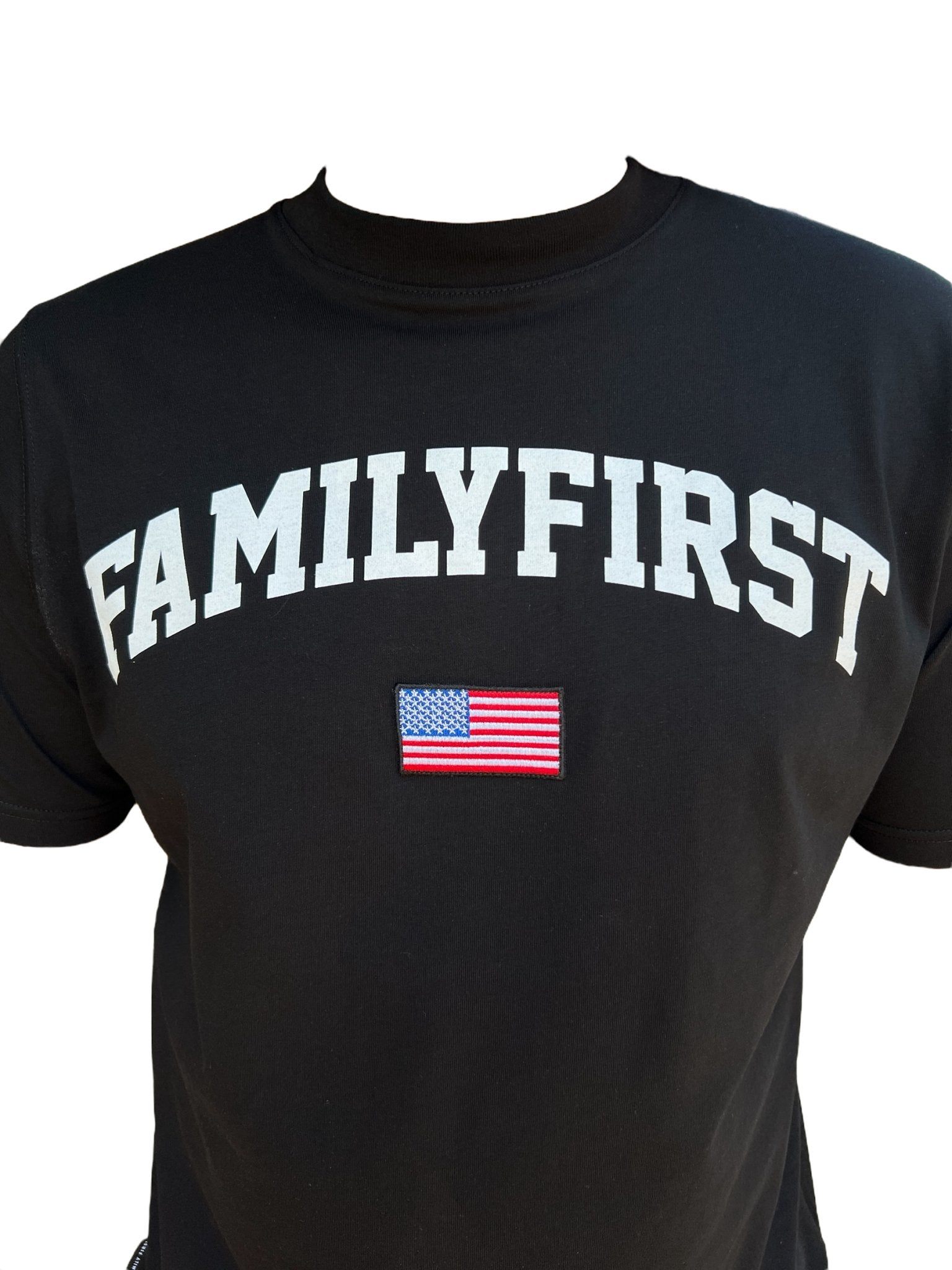 The Family First TS2416 T-Shirt College Black by FAMILY FIRST is made from 100% cotton and showcases "FAMILYFIRST" prominently in white, accompanied by a small American flag patch beneath.