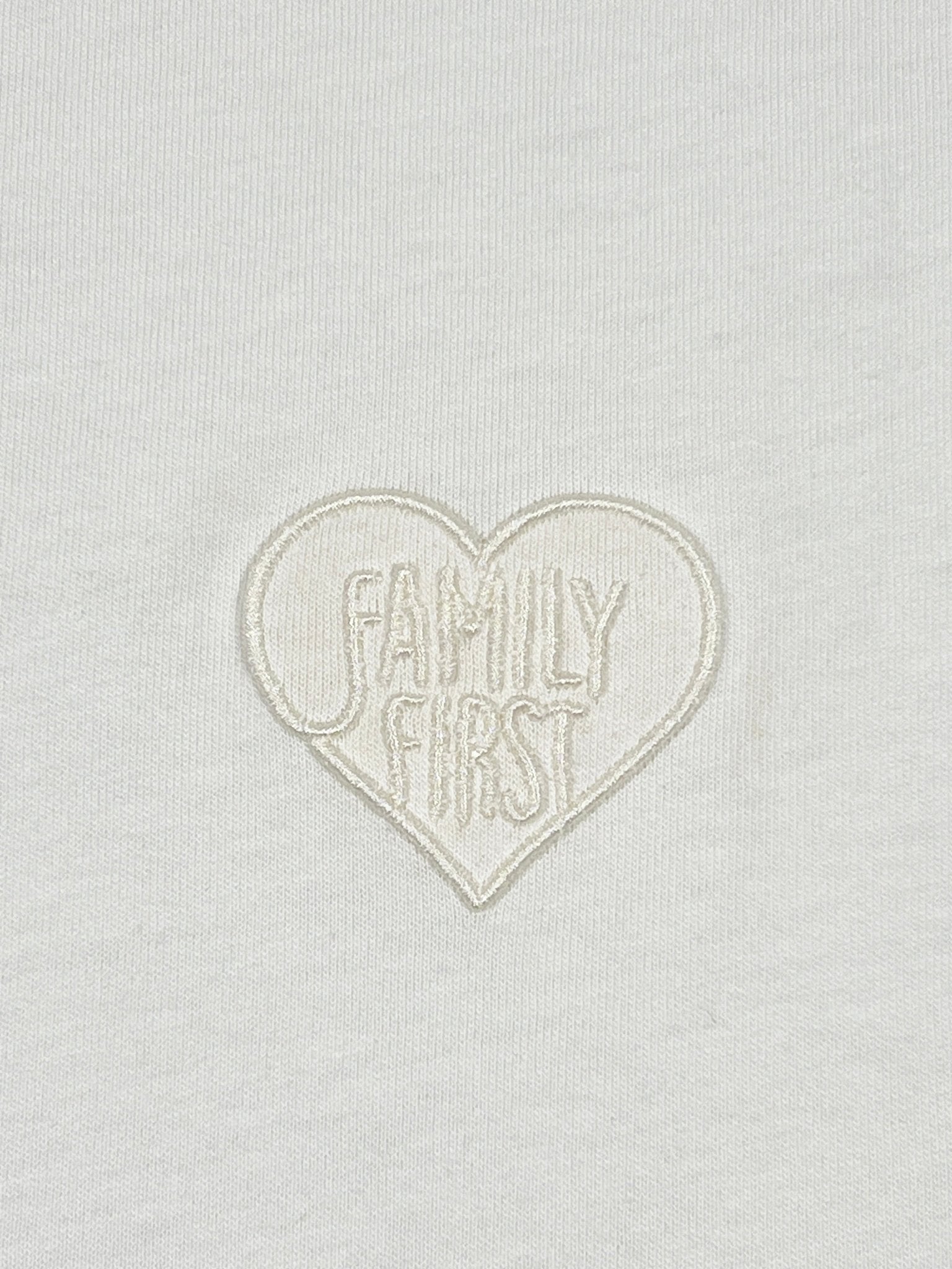 The Family First TS2504 T-shirt in Heart Tone White showcases a heart-shaped embroidered logo displaying "FAMILY FIRST," perfect for those who cherish unity, embodying the brand's essence.