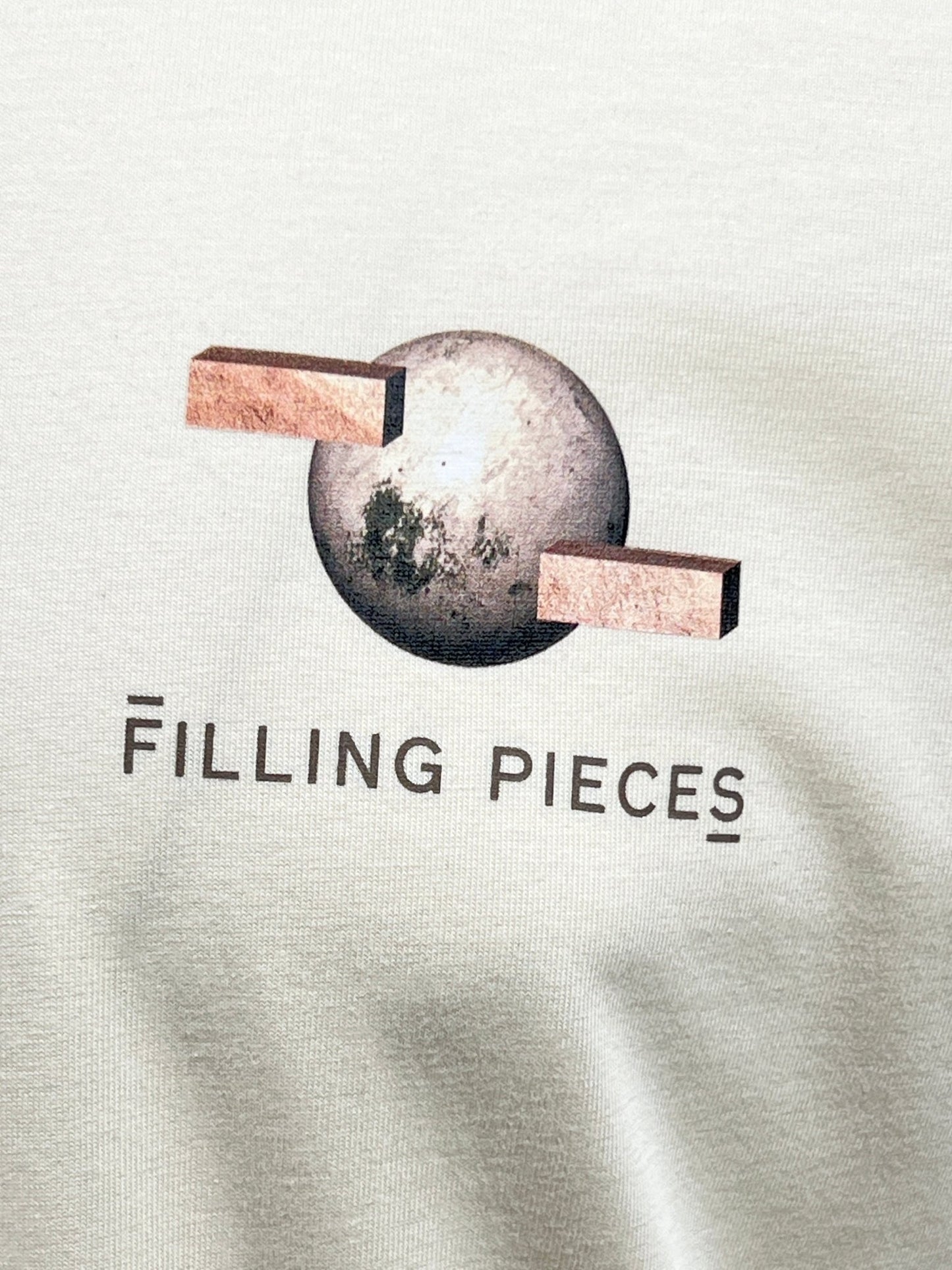 Close-up of a graphic on the FILLING PIECES BALANCE ANTIQUE WHITE T-SHIRT, featuring a sphere intersected by two wooden blocks with the text "FILLING PIECES" beneath it, adding to the T-shirt's unique backprint.