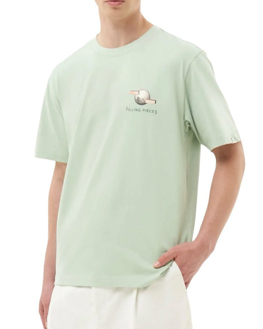 A person wearing a FILLING PIECES BALANCE frosty green T-shirt made of organic cotton, featuring a small graphic and the text "Filling Pieces" on the chest, paired with white pants. Hand in pocket.