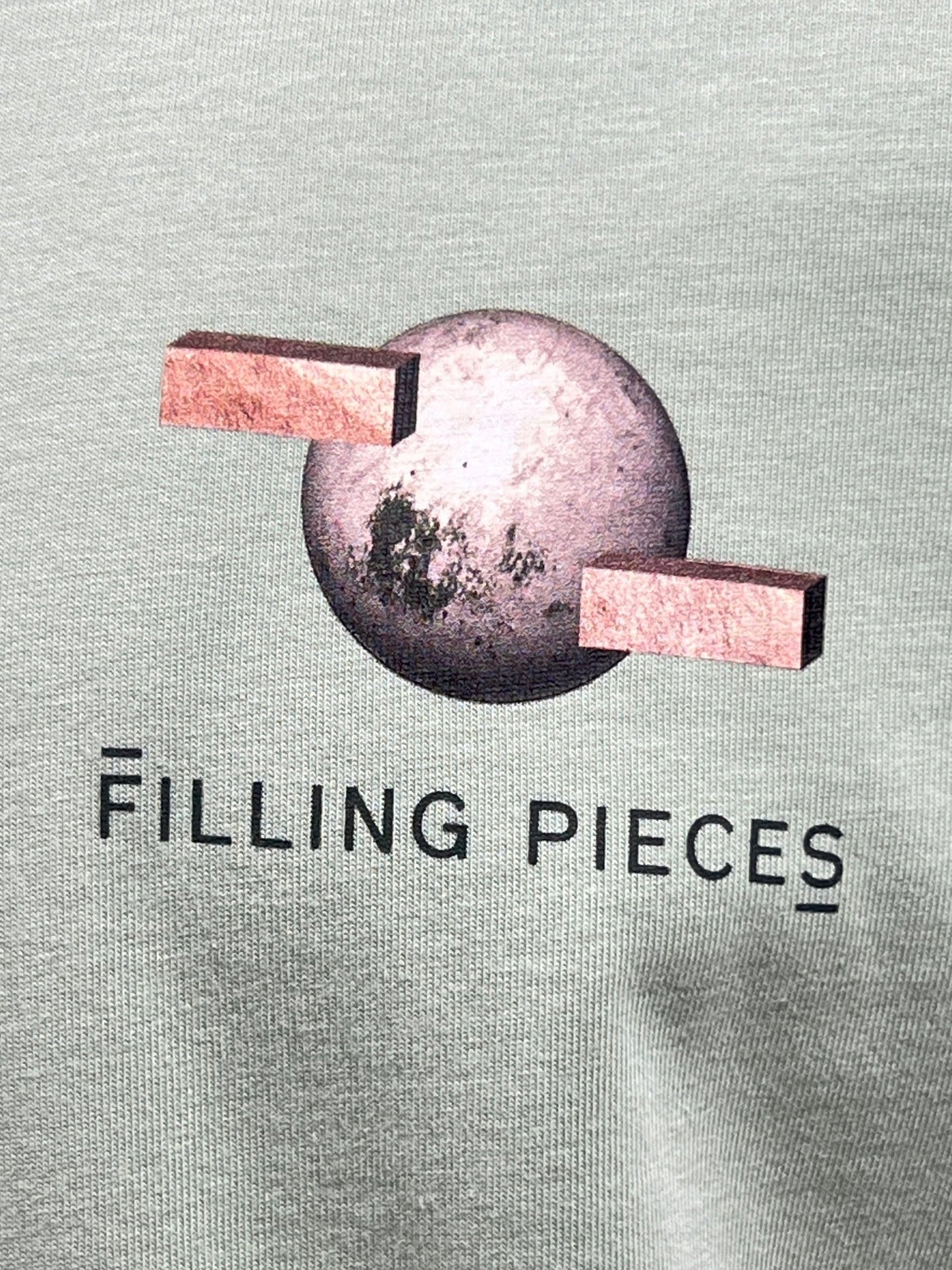 Close-up of a design on organic cotton fabric featuring a textured sphere with two rectangular shapes extending from it, above the text "FILLING PIECES," showcasing the FP logo on the FILLING PIECES BALANCE FROSTY GREEN T-SHIRT.