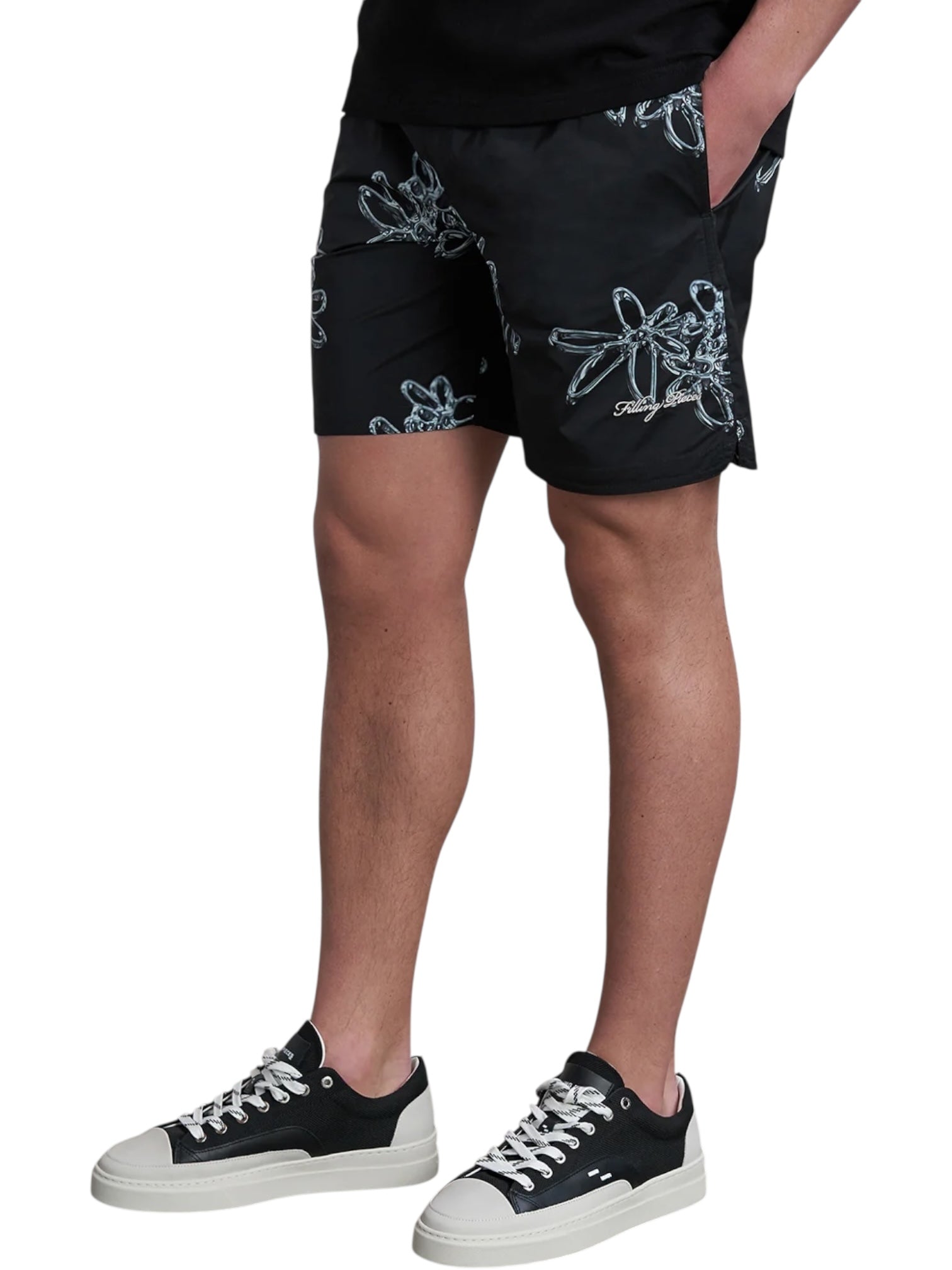 A person wearing FILLING PIECES Chrome Swimshorts Black, a black shirt, and black-and-white sneakers stands with one hand in their pocket.