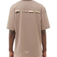 A person with dark braided hair wearing a FILLING PIECES CONSTRUCT FOSSIL T-SHIRT from FILLING PIECES, made of organic cotton in beige, featuring a printed graphic and the text "BRIDGE THE GAP" on the back.