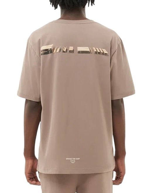 A person with dark braided hair wearing a FILLING PIECES CONSTRUCT FOSSIL T-SHIRT from FILLING PIECES, made of organic cotton in beige, featuring a printed graphic and the text "BRIDGE THE GAP" on the back.