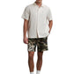 A man stands against a white background wearing a short-sleeve beige shirt, FILLING PIECES Flower Shorts Camo featuring white graffiti, white socks, and black loafers.