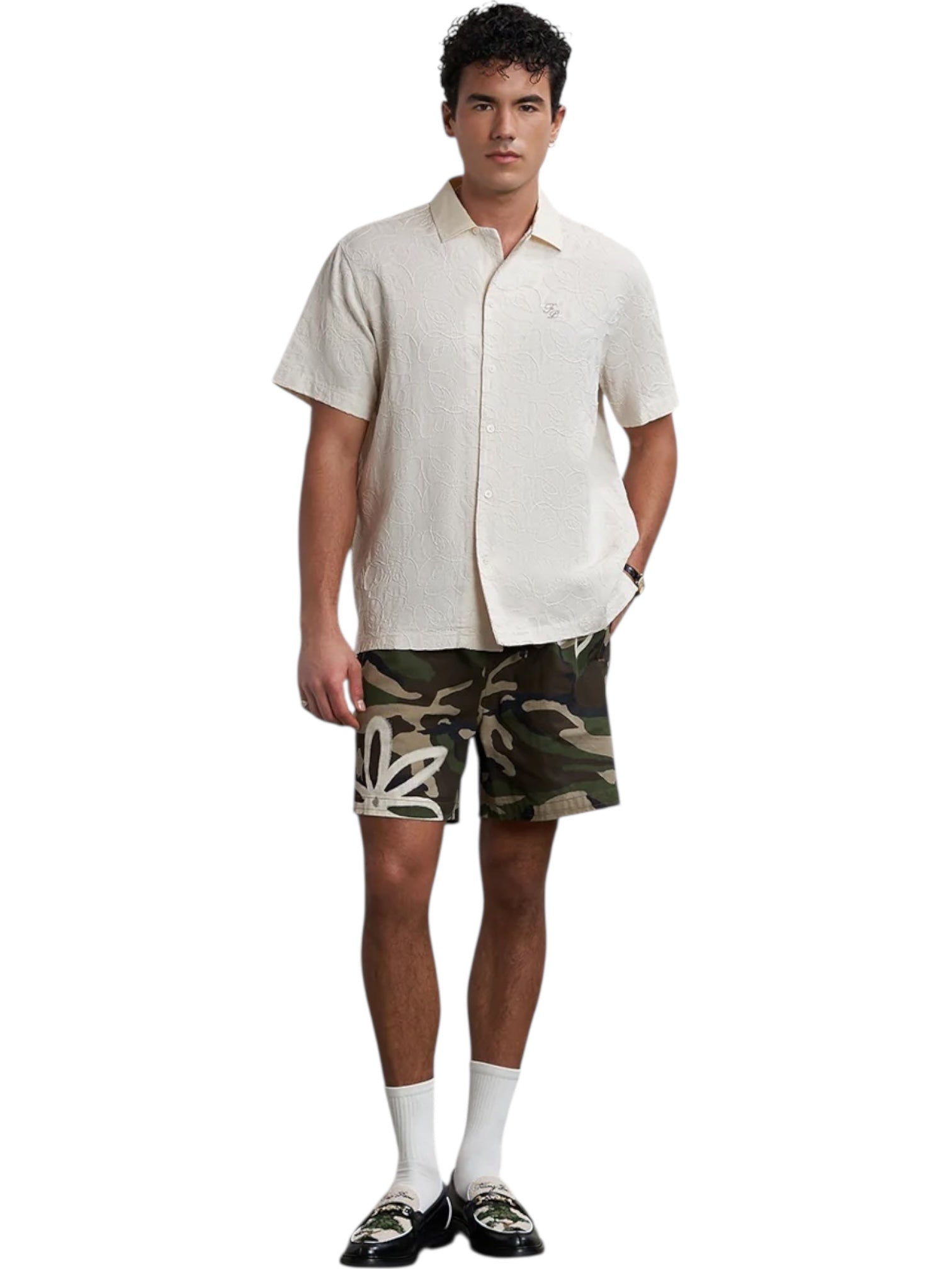 A man stands against a white background wearing a short-sleeve beige shirt, FILLING PIECES Flower Shorts Camo featuring white graffiti, white socks, and black loafers.