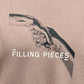 Close-up of the FILLING PIECES HANDSHAKE FOSSIL T-SHIRT, showcasing an image of two hands clasping each other with the text "FILLING PIECES" printed beneath the image.