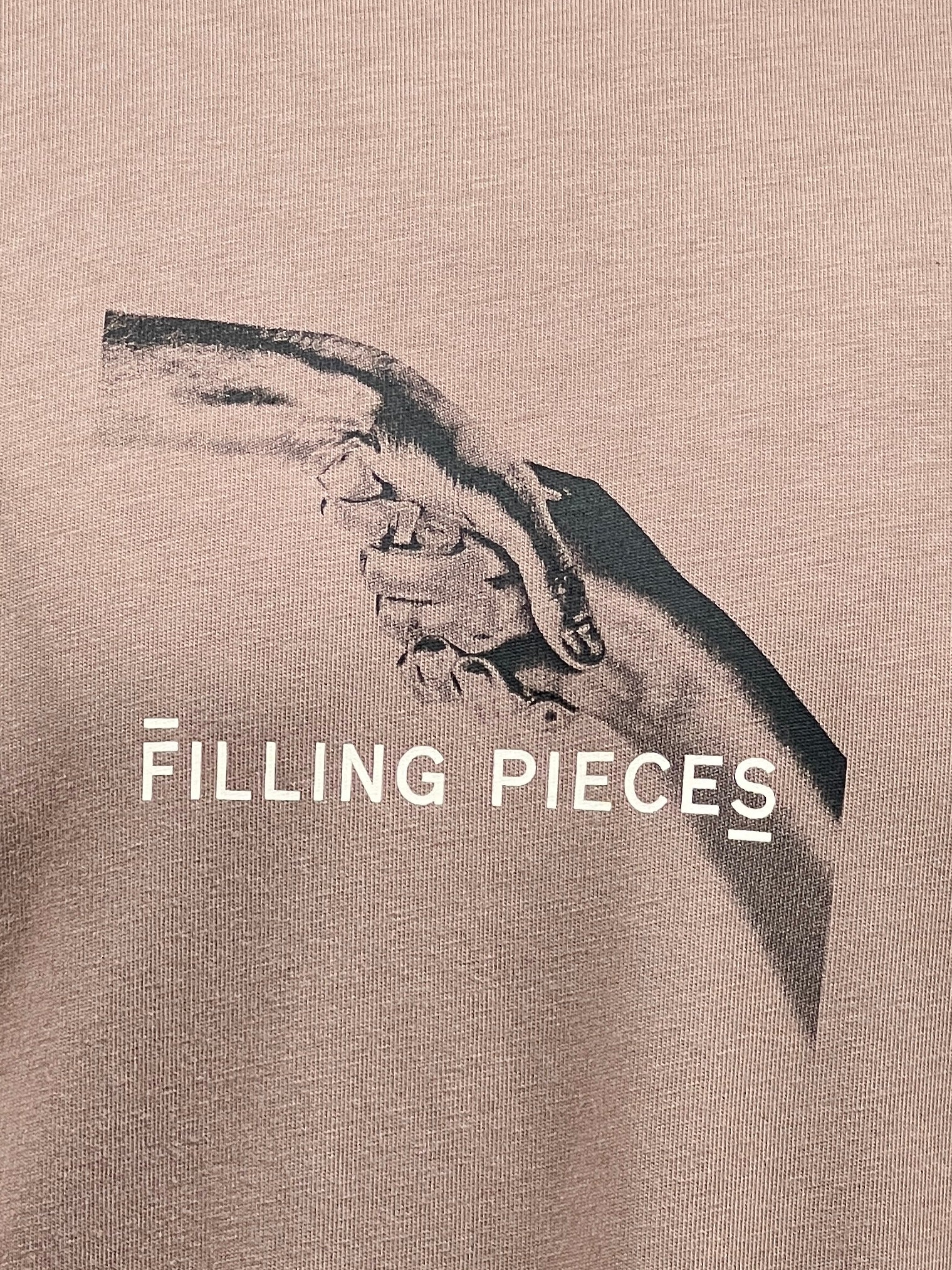 Close-up of the FILLING PIECES HANDSHAKE FOSSIL T-SHIRT, showcasing an image of two hands clasping each other with the text "FILLING PIECES" printed beneath the image.