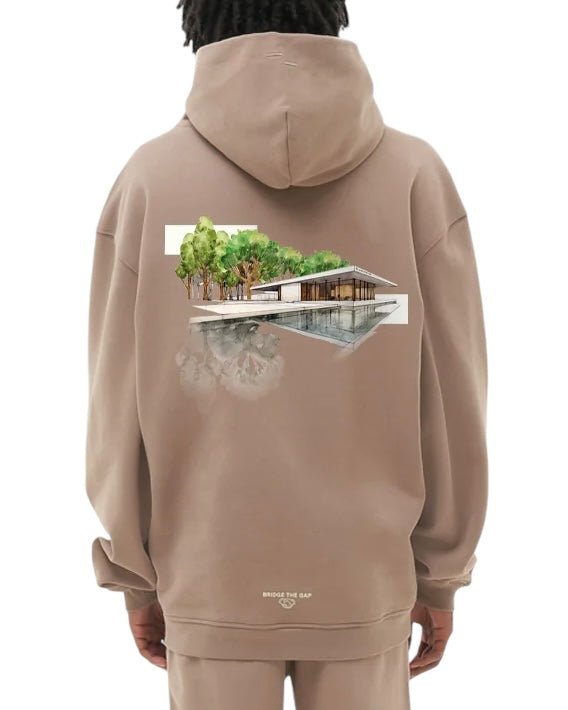A person in the Filling Pieces Hoodie Pavilion Fossil by FILLING PIECES displays a modern house and trees illustration on the back of their beige organic cotton hoodie.